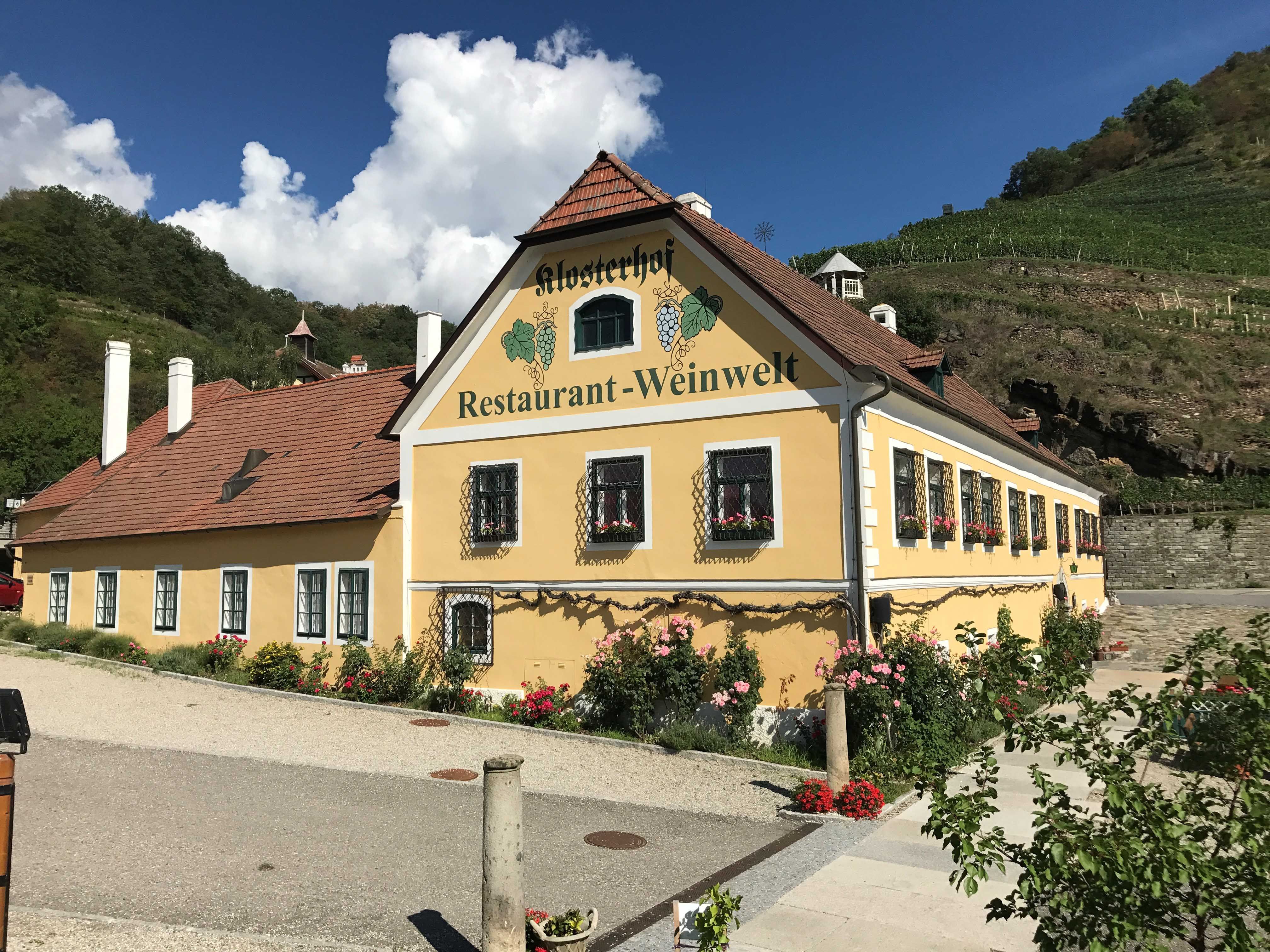 A "Klosterhof Restaurant-Weinwelt" feliratú sárga épület külső nézete, virágokkal és szőlőültetvényekkel körülvéve.