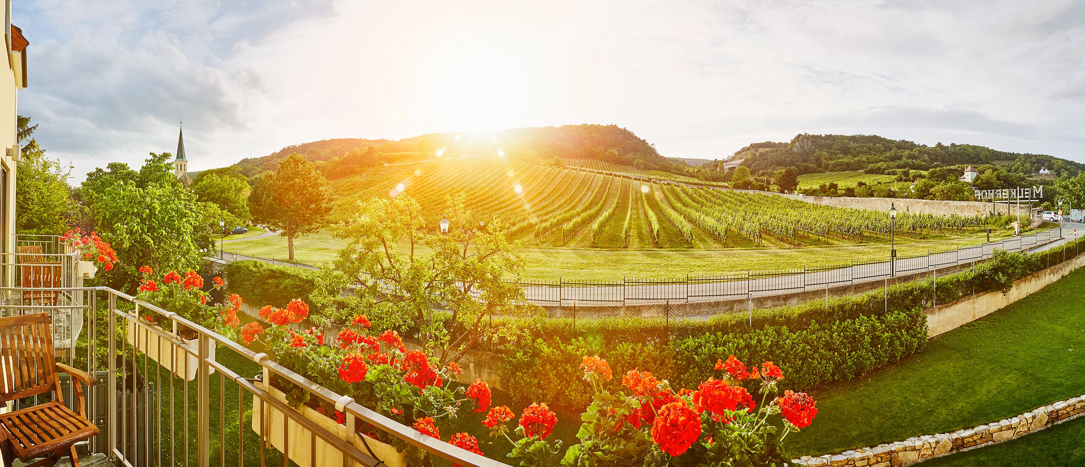 Die sanften Hügel der Weinregion laden zu einem entspannten Aufenthalt ein, während die warmen Sonnenstrahlen die Weinreben in goldenes Licht tauchen. Bunte Geranien blühen auf dem Balkon und verleihen der Szenerie einen Hauch von Gemütlichkeit und Lebensfreude.