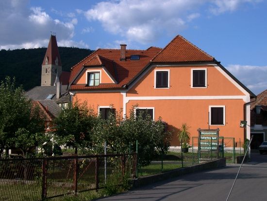 Narancssárga színű ház, vörös tetőcserepekkel, a háttérben egy templomtorony.