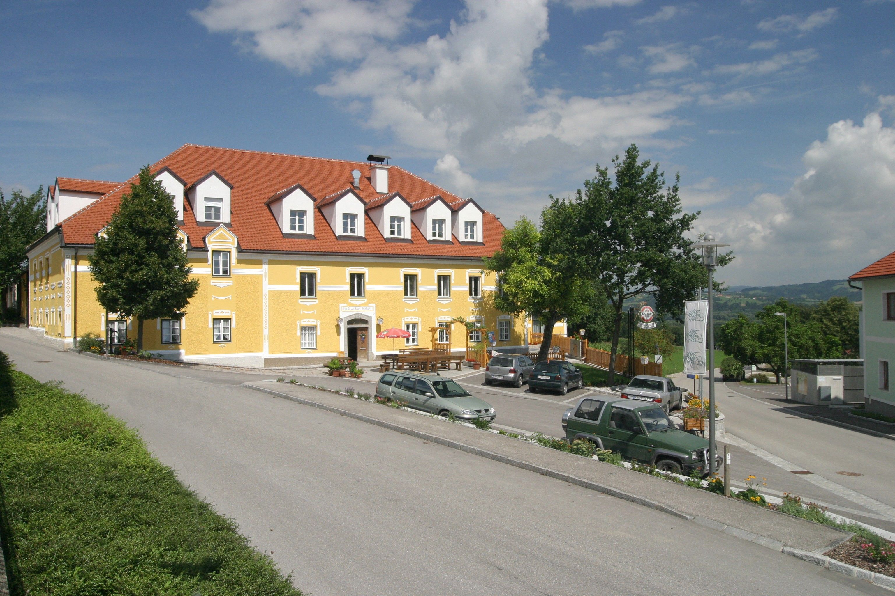 Egy sárga épület piros tetővel, autókkal és fákkal körülvéve, kék ég alatt.