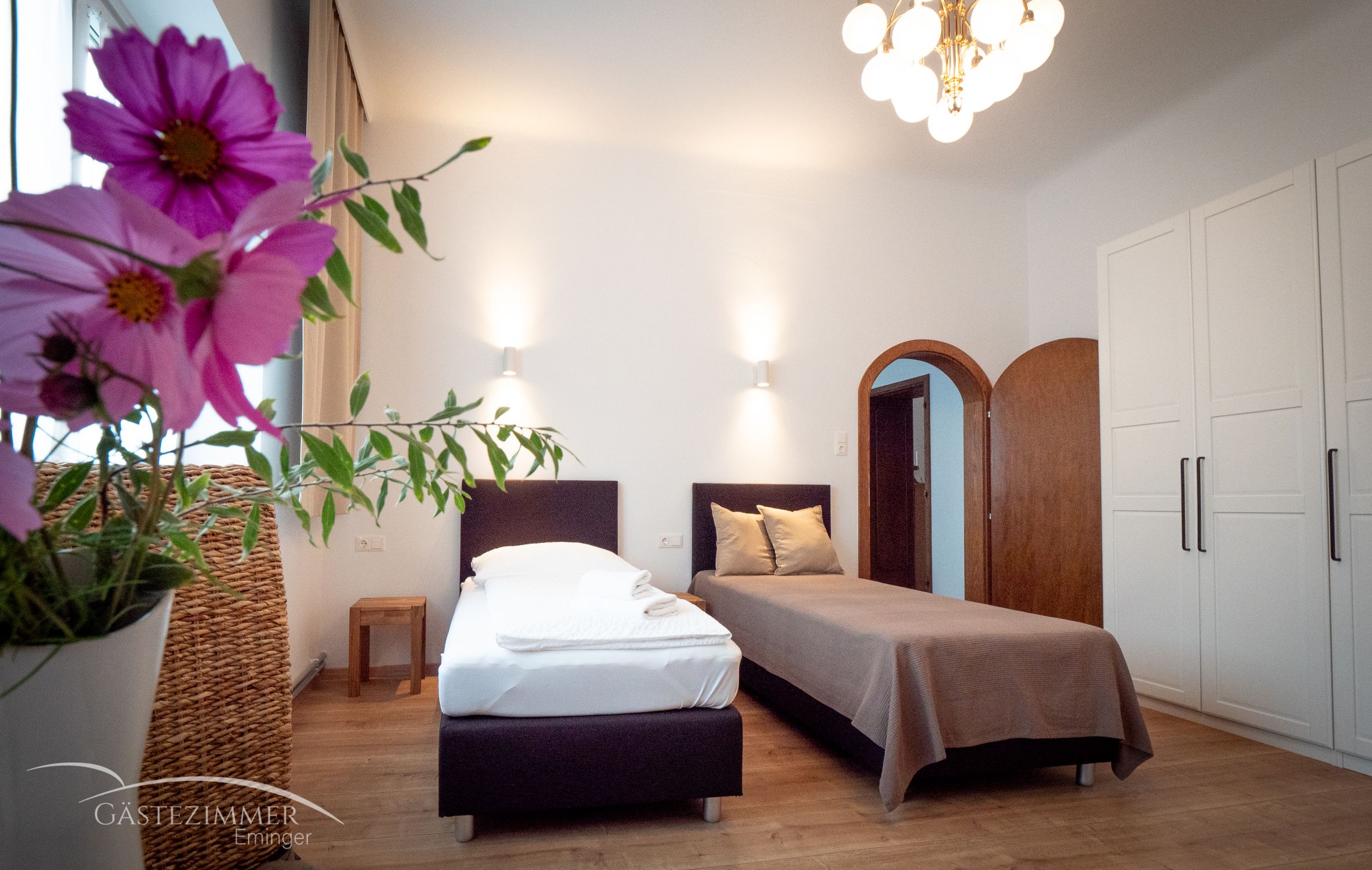 Hangulatos kétágyas szoba, virágokkal az előtérben, két ággyal, szekrénnyel és lámpával a mennyezeten.