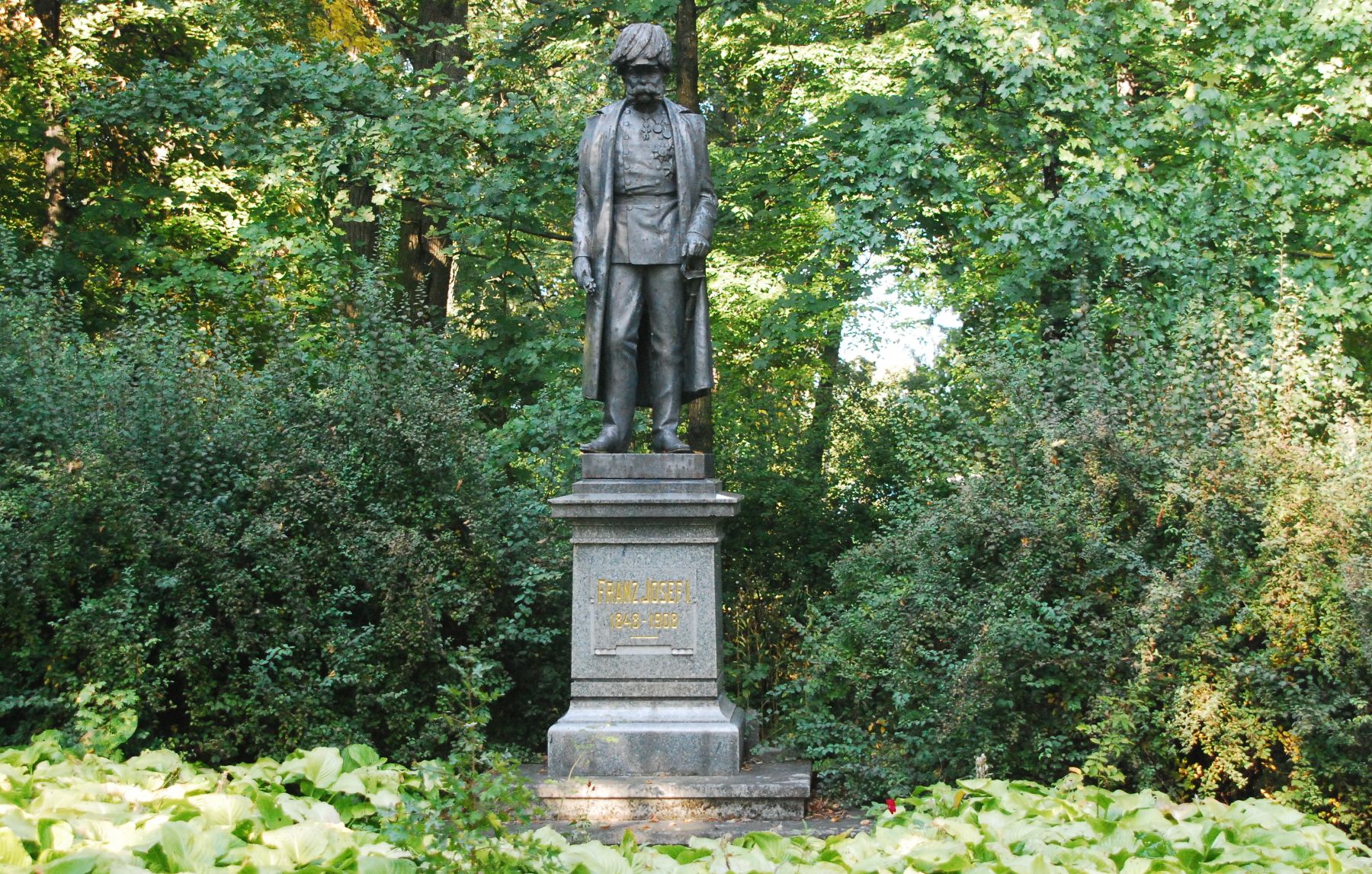 I. Ferenc József császár szobra egy parkban, fákkal és cserjékkel körülvéve.