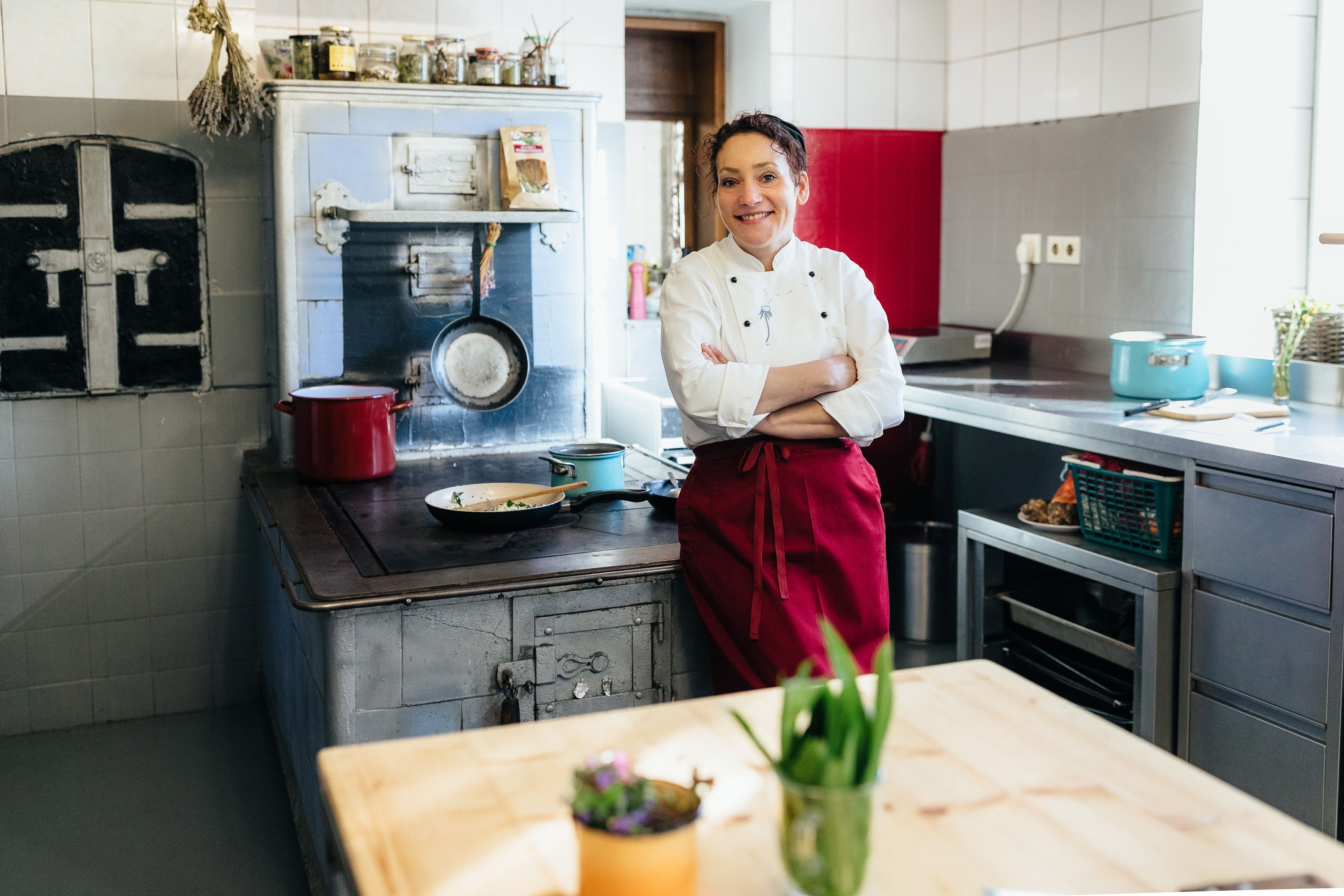 Egy szakács fehér kabátban és piros kötényben áll a konyhában egy régi tűzhely előtt.