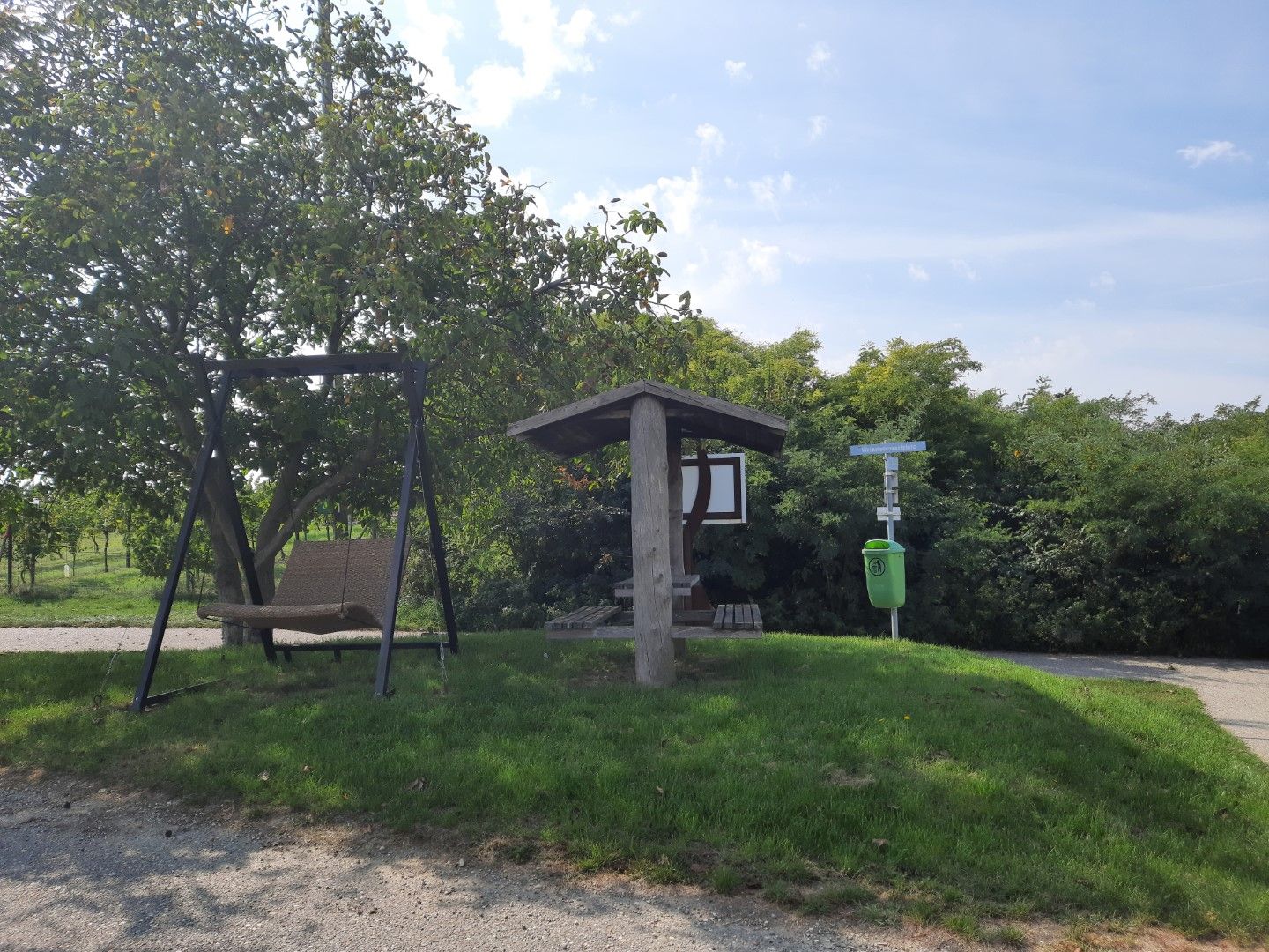 Pihenőhely hintával és fedett ülőhely egy fa és egy szemetes kuka mellett a réten.