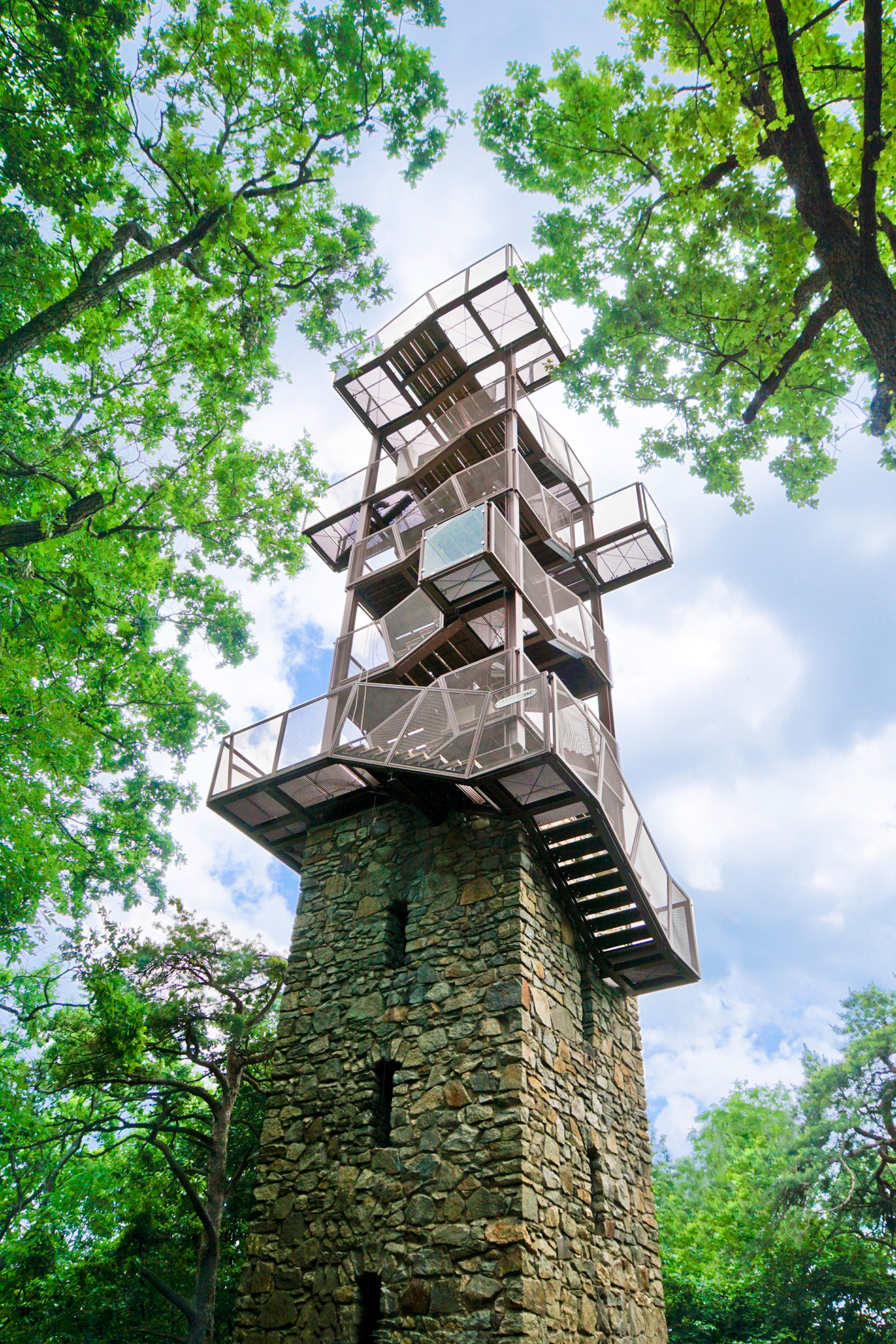 A kék égbe egy modern, fémből és kőből készült, zöld fákkal körülvett kilátótorony emelkedik.