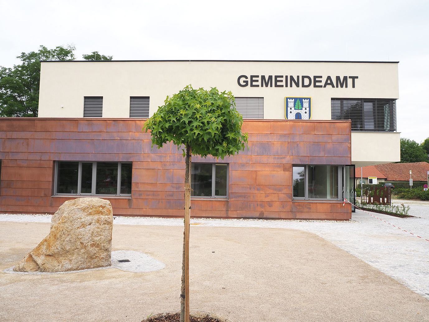 A Sieghartskircheni önkormányzati hivatal modern épülete, előtérben fa és kő.