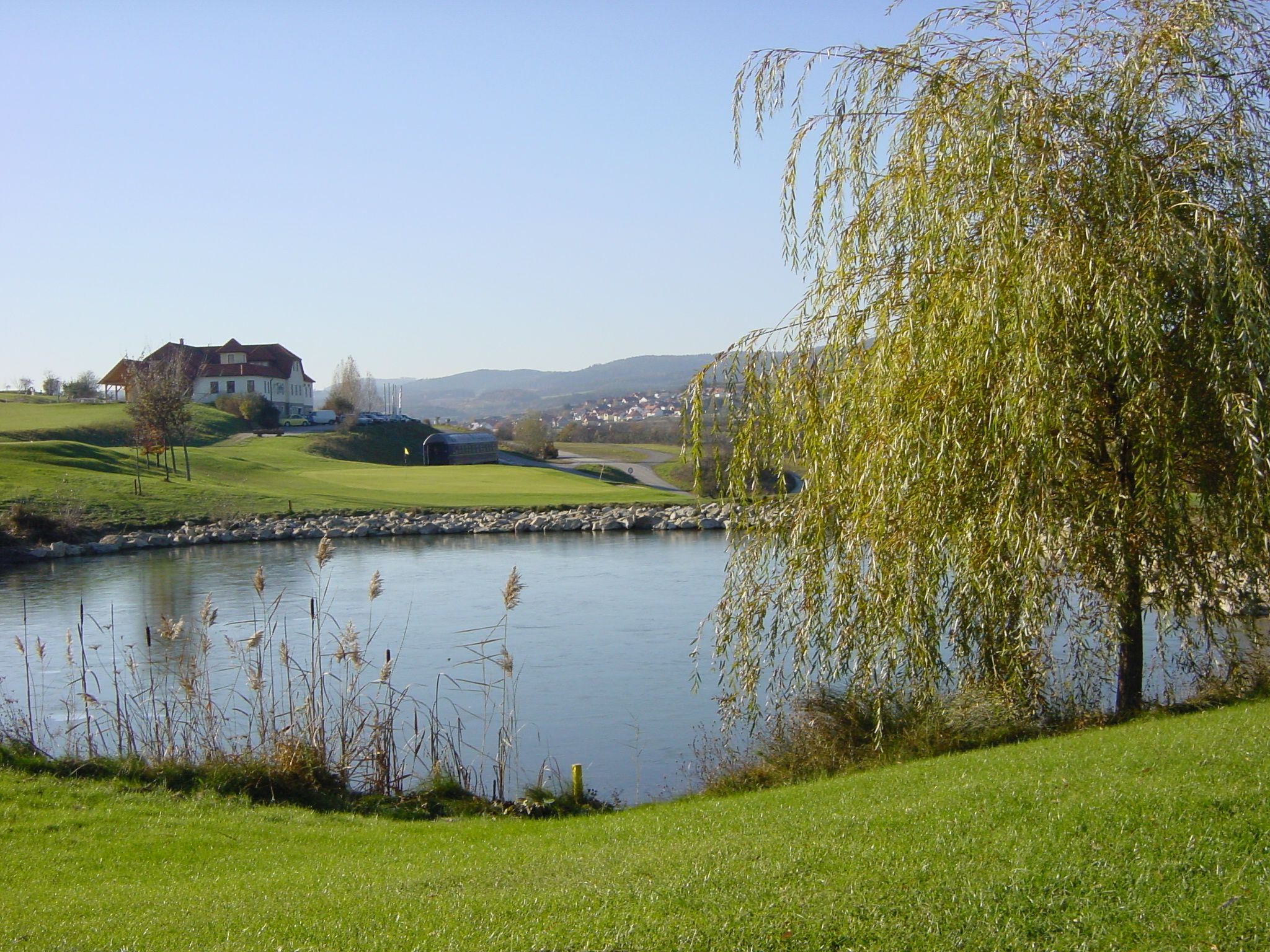 Tájkép golfpályával, tóval és épületekkel a háttérben.