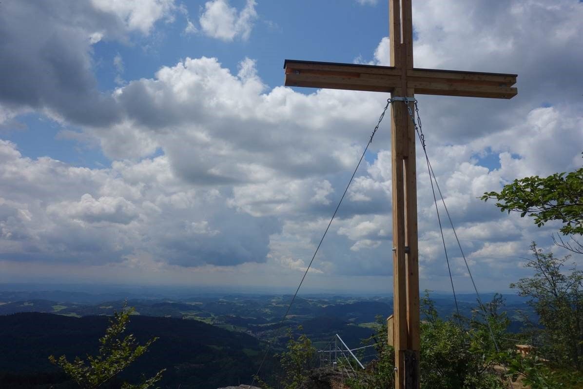Csúcskereszt a Burgsteinmauer kilátóhegyen, kilátással a felhős égre és a tájra.