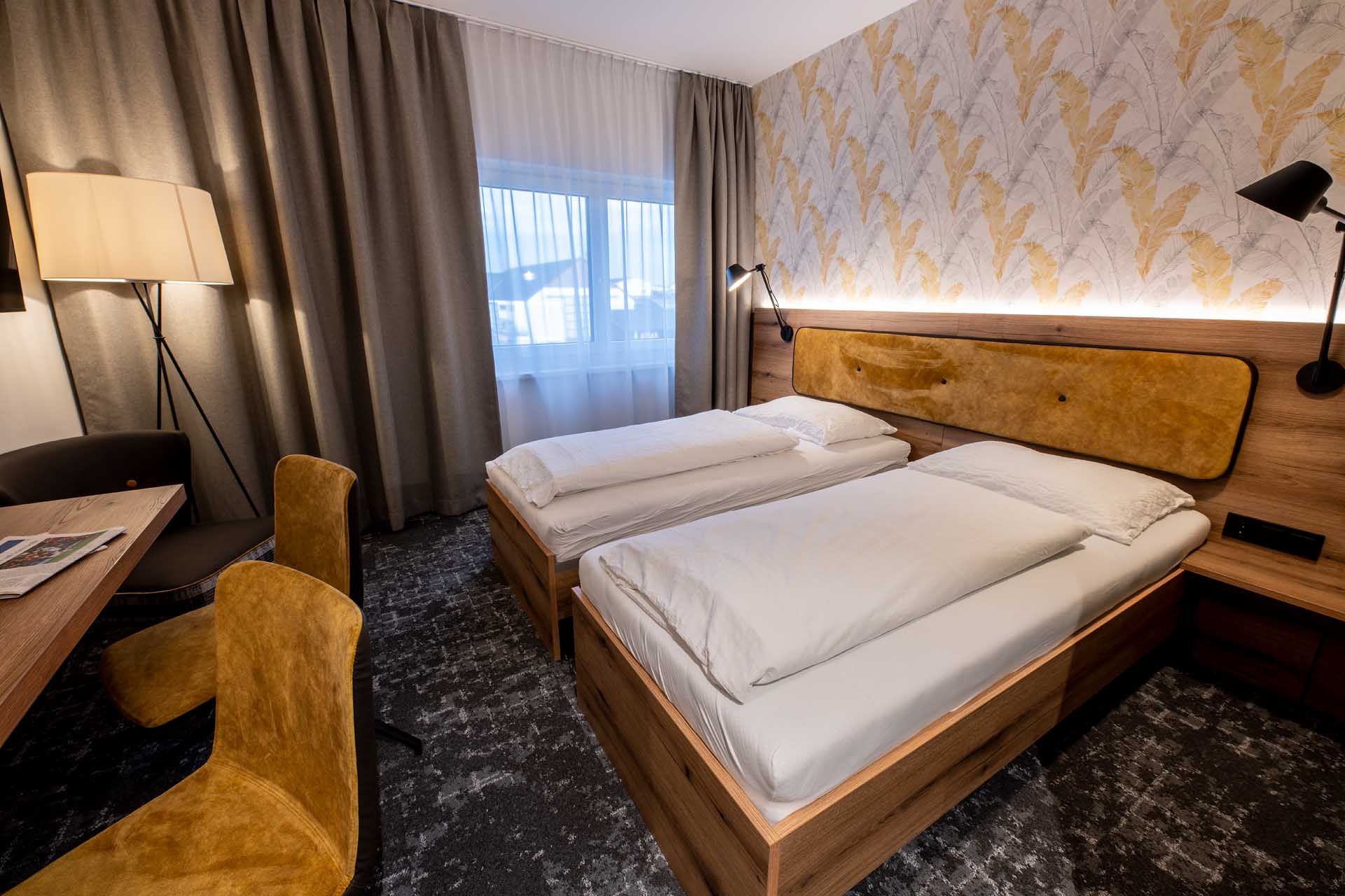 Modern vendégszoba két egyszemélyes ággyal, fabútorokkal, sárga székekkel és állólámpával. A falon világos színű függönyök és mintás tapéta.