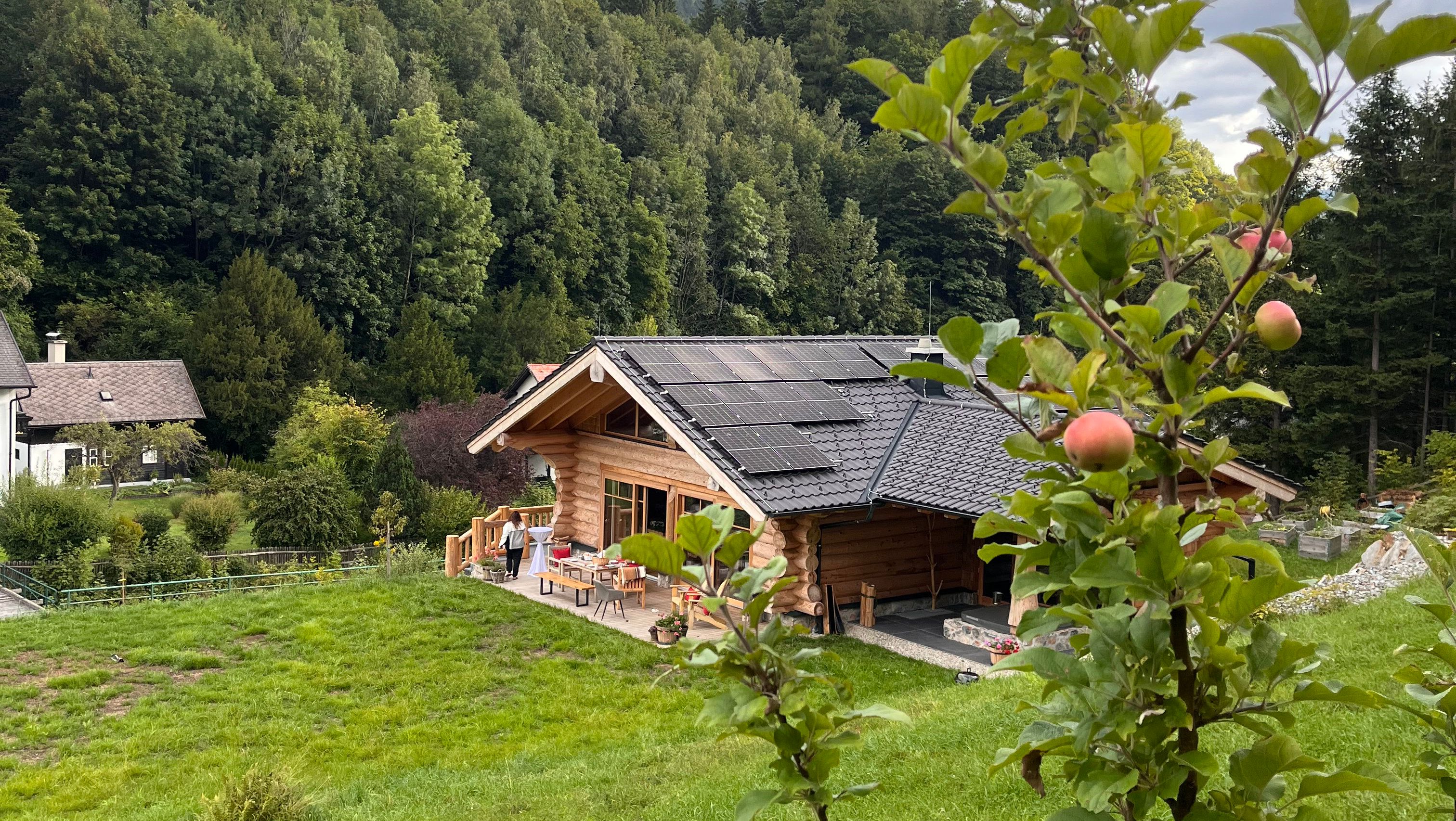 Fából készült faház napelemekkel a tetőn, zöld vidékkel és fákkal körülvéve. Az előtérben egy almafa érett almákkal.