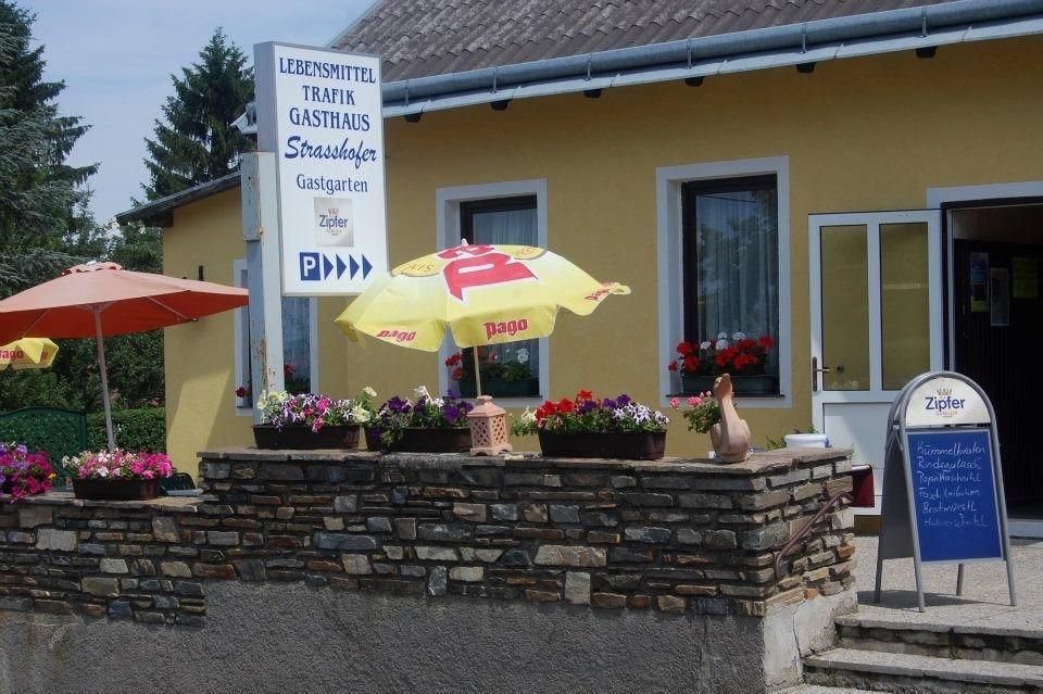 Egy sárga épület külső nézete, élelmiszer, dohánybolt és vendéglő táblákkal. Az épület előtt napernyők és virágok.