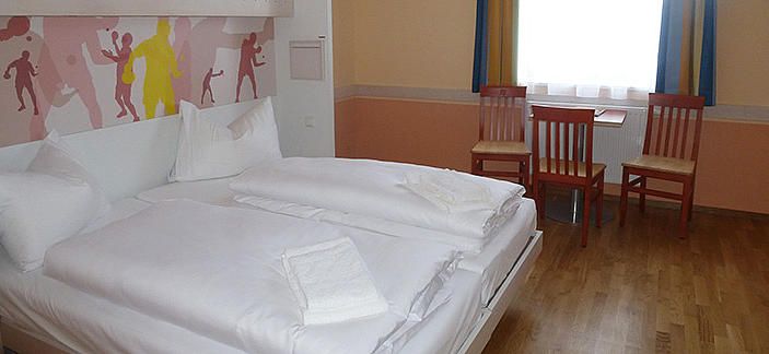 Hotelszoba franciaággyal, két székkel és egy asztallal az ablak előtt.