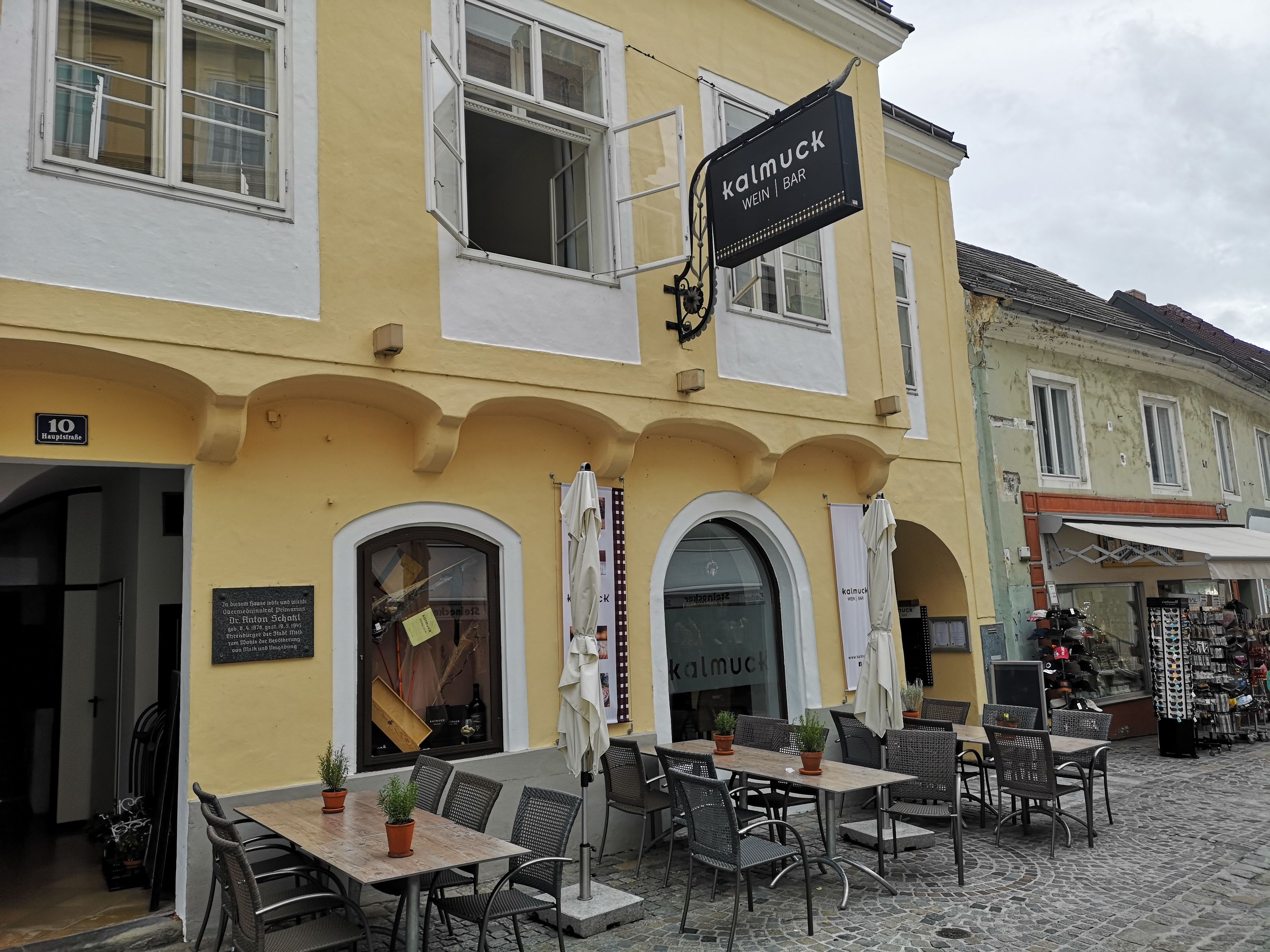 Egy sárga épület külső nézete a "Kalmuck Wine Bar" felirattal. Az épület előtt asztalok és székek állnak.