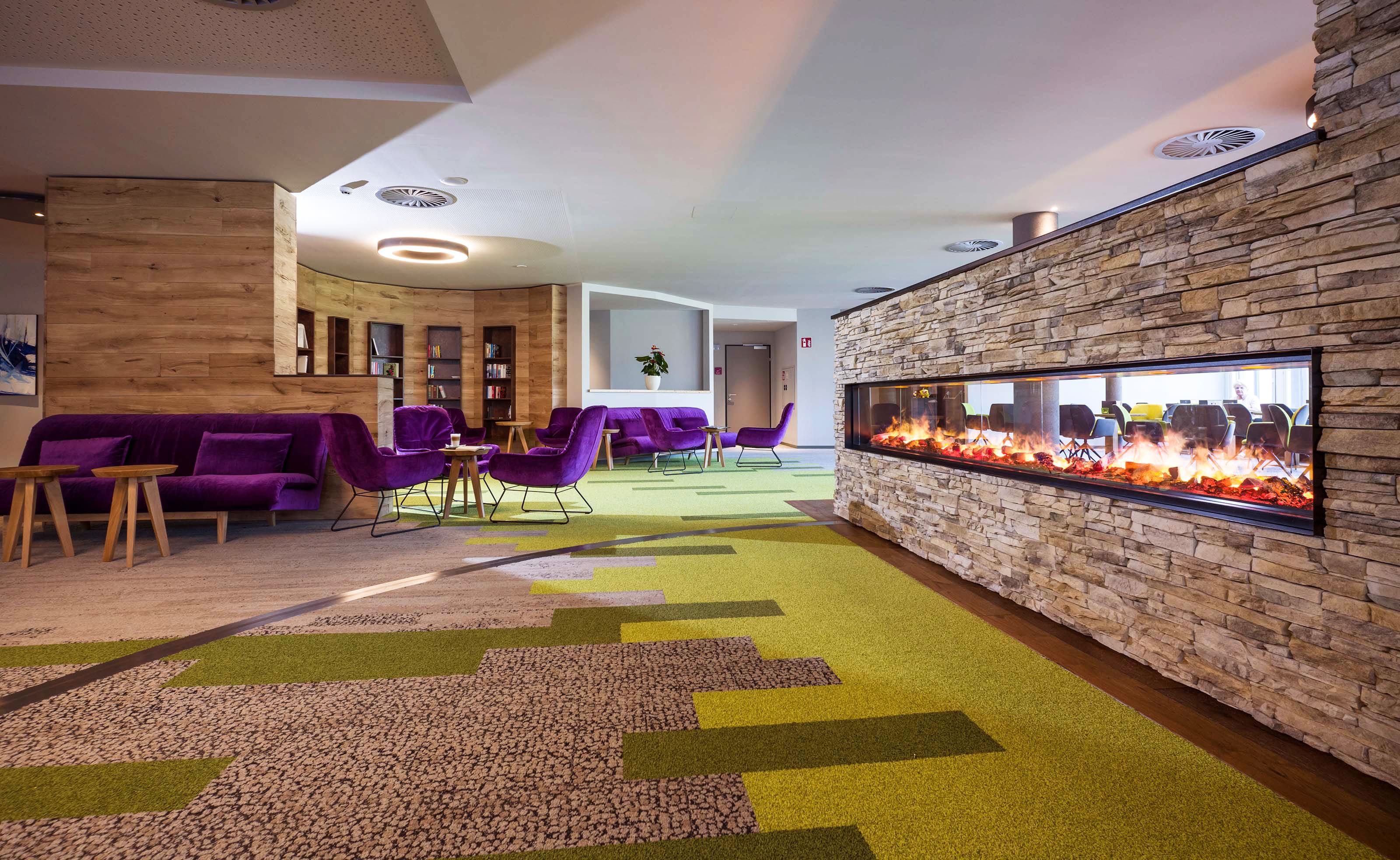 Modern szálloda lobby zöld-barna szőnyeggel, kandallóval, lila kanapékkal és székekkel, valamint faasztalokkal.