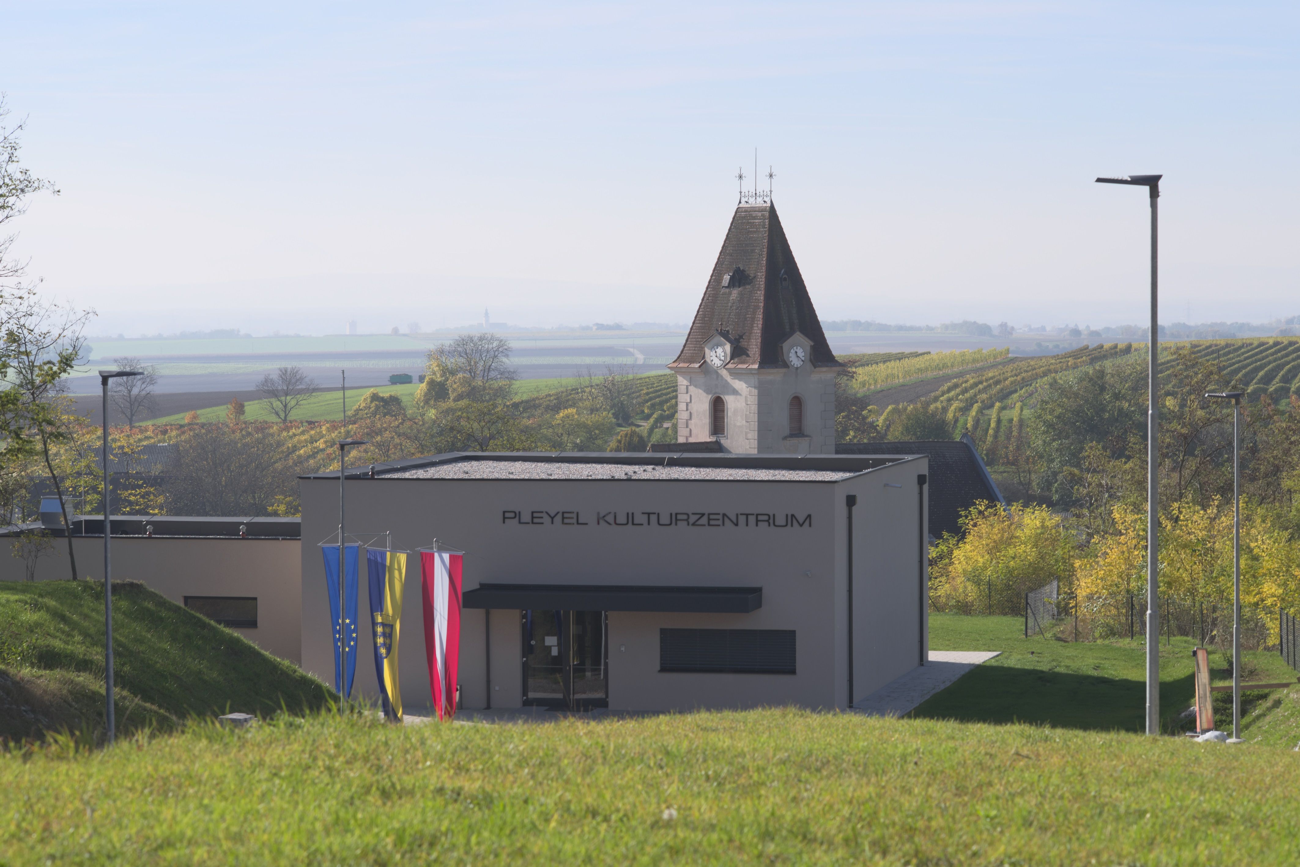 A "Pleyel Kulturális Központ" feliratú modern épület egy vidéki tájkép előtt, szőlőültetvényekkel és egy templomtoronnyal.