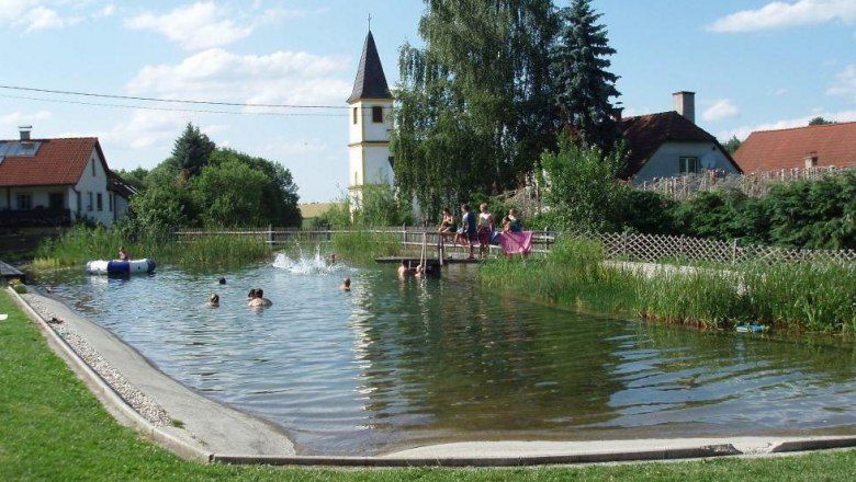 Természetes fürdőző tó, ahol emberek úsznak. A háttérben templomtorony és házak láthatók.