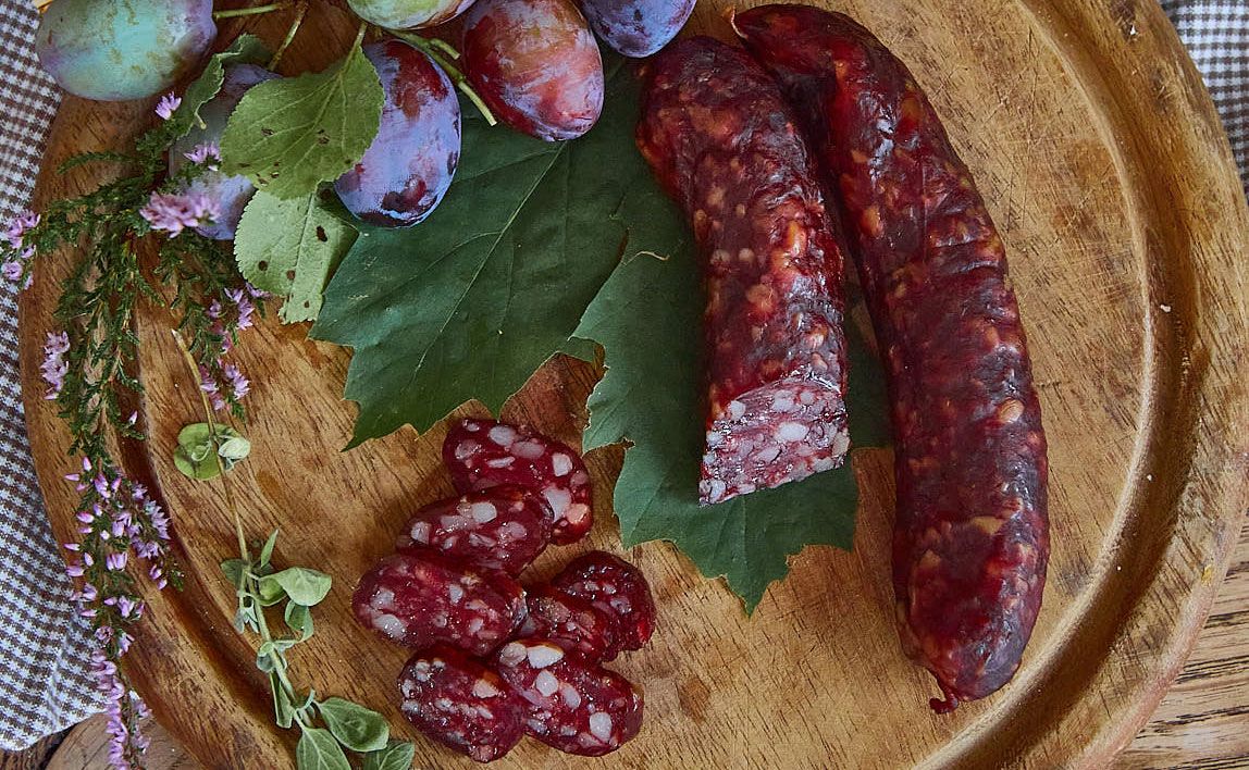 Kaminwurzn és szilva zöld levelekkel és fűszernövényekkel díszített fatáblán.