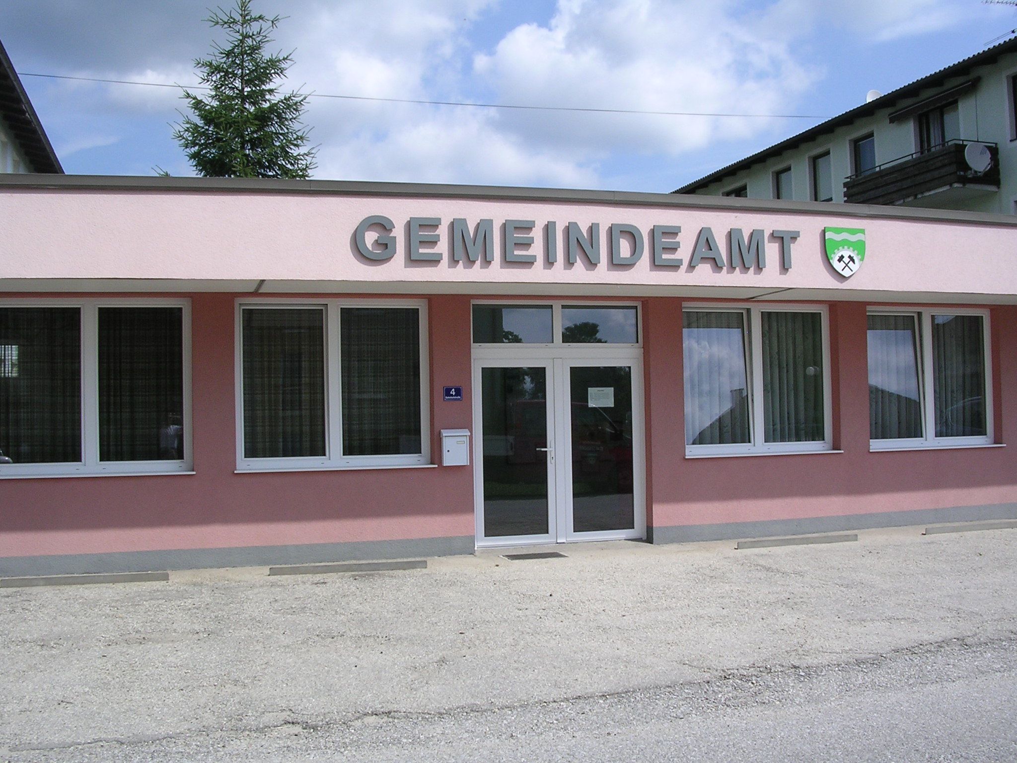 Egy rózsaszínű épület külső nézete "GEMEINDEAMT" felirattal és címerrel.
