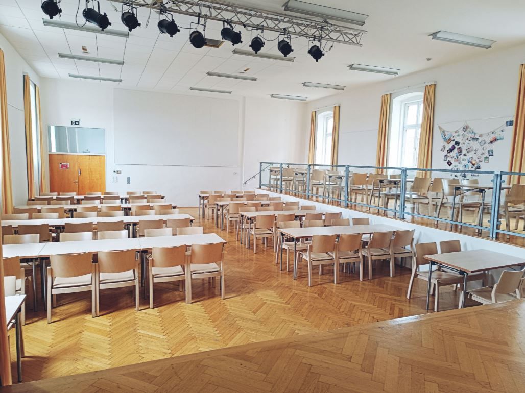 Egy üres terem, fapadlóval, asztalokkal és székekkel, színpaddal és világítással a mennyezeten.