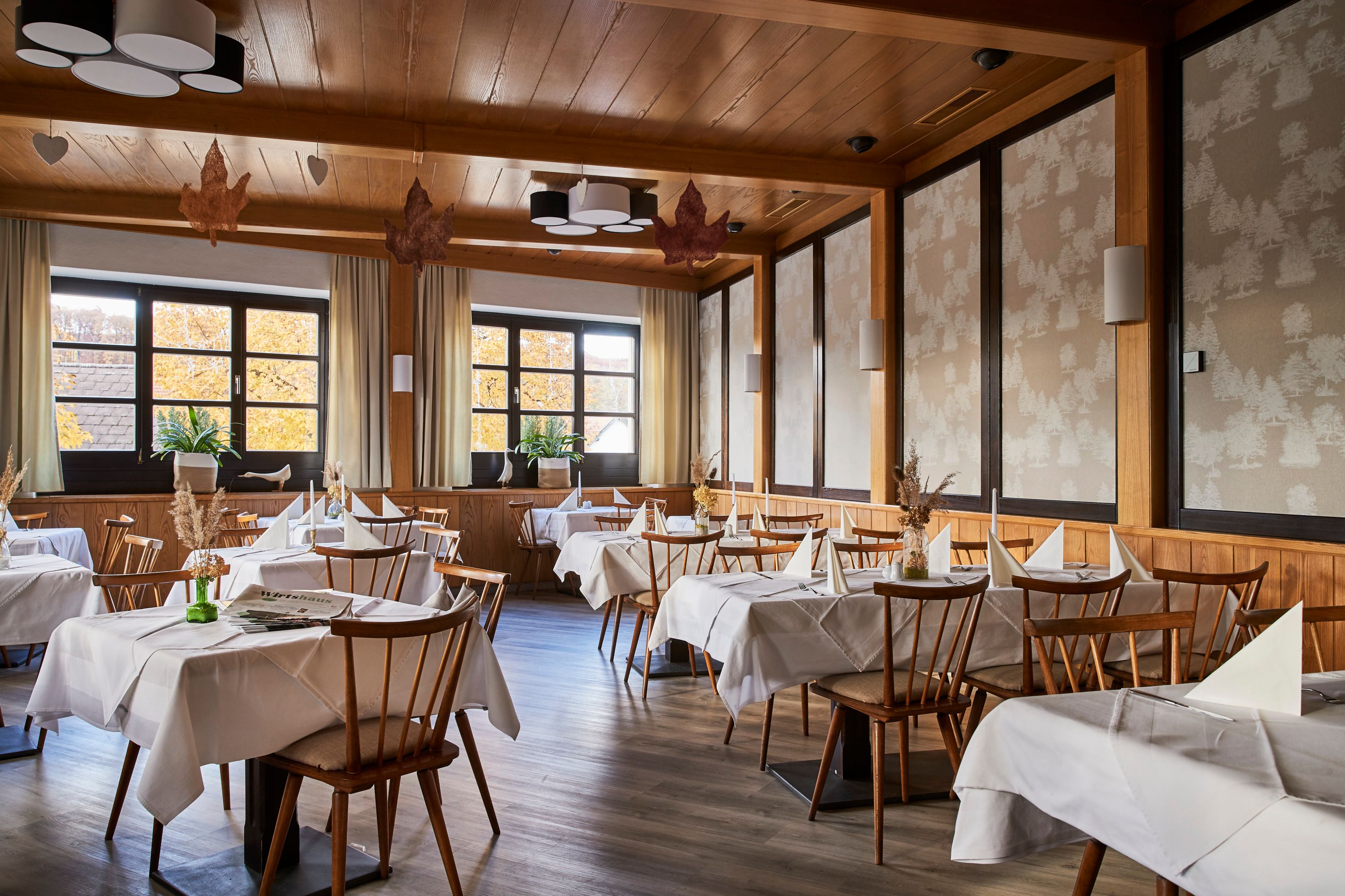 Elegáns étterem, fából készült bútorokkal, fehér terítőkkel és nagy ablakokkal, amelyeken őszi fák láthatók.