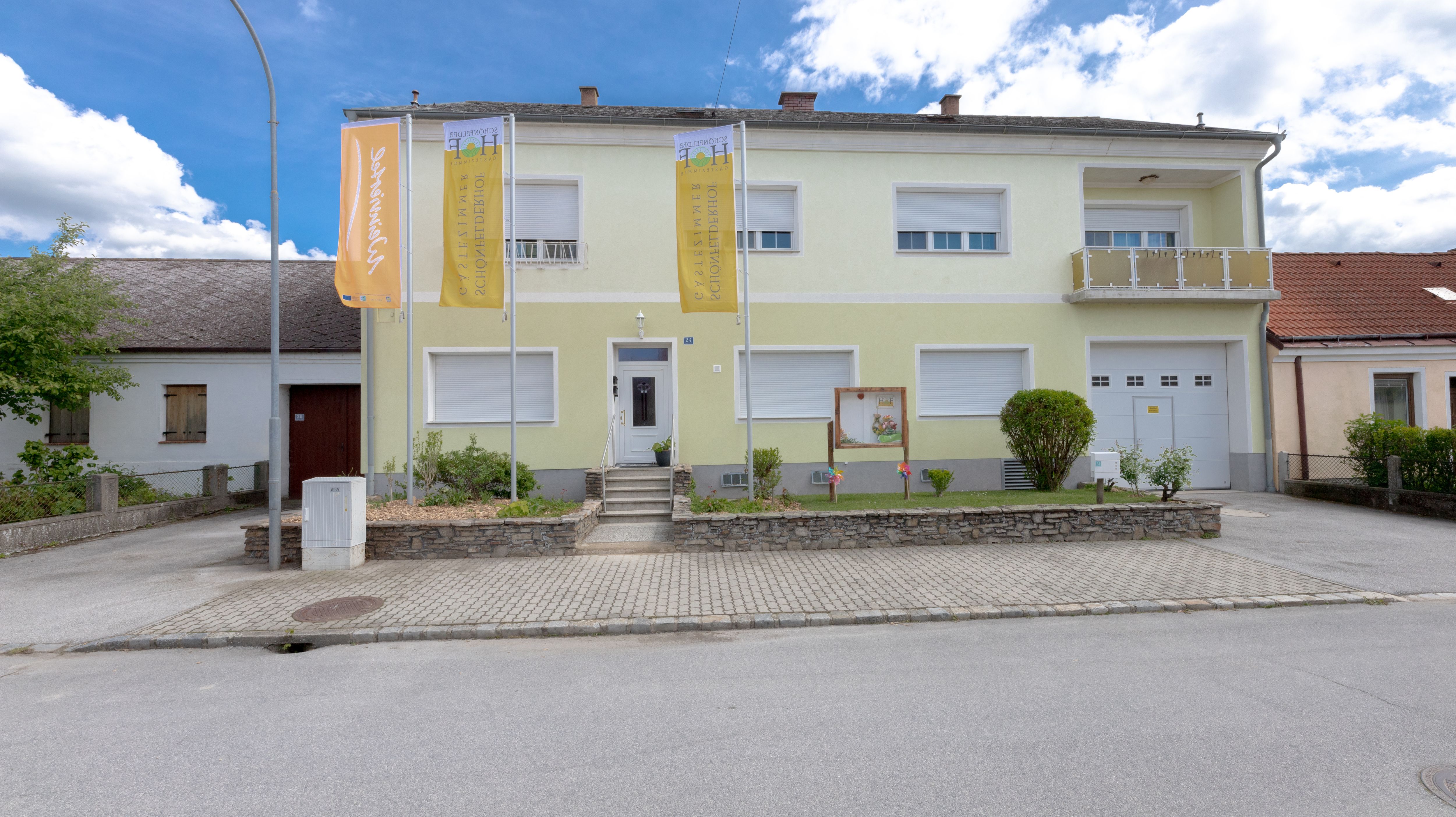 Egy kétszintes épület külső nézete, sárga homlokzattal, előtte két zászlóval.