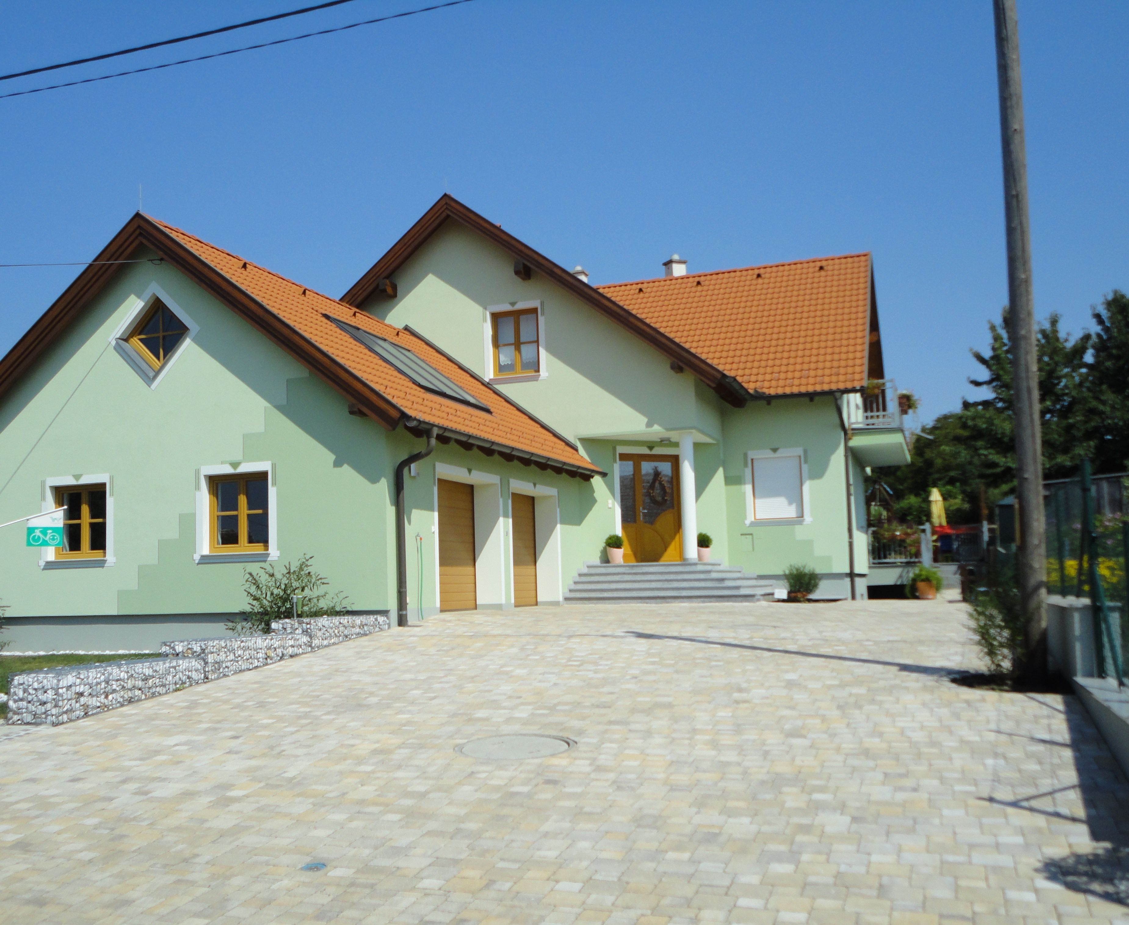 Modern, kétszintes, zöldre festett, piros tetőcseréppel fedett ház, amelyet egy kövezett udvar vesz körül.