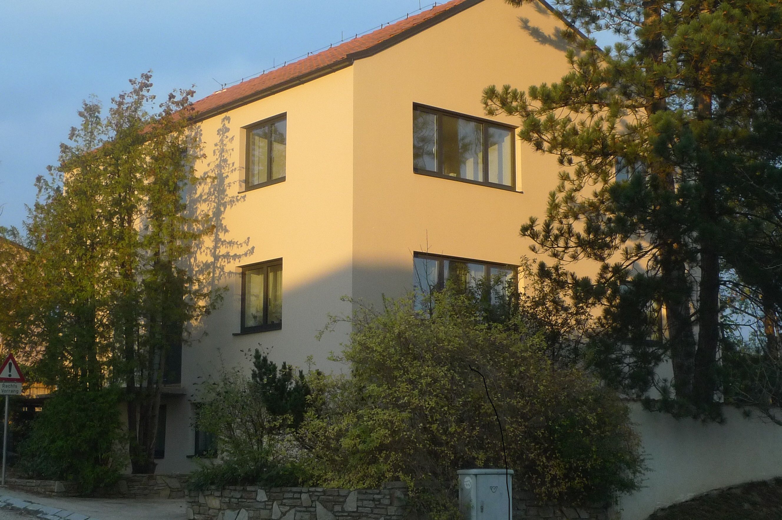 Modern, háromszintes ház, sárga homlokzattal és piros tetőcseréppel, fákkal és bokrokkal körülvéve.