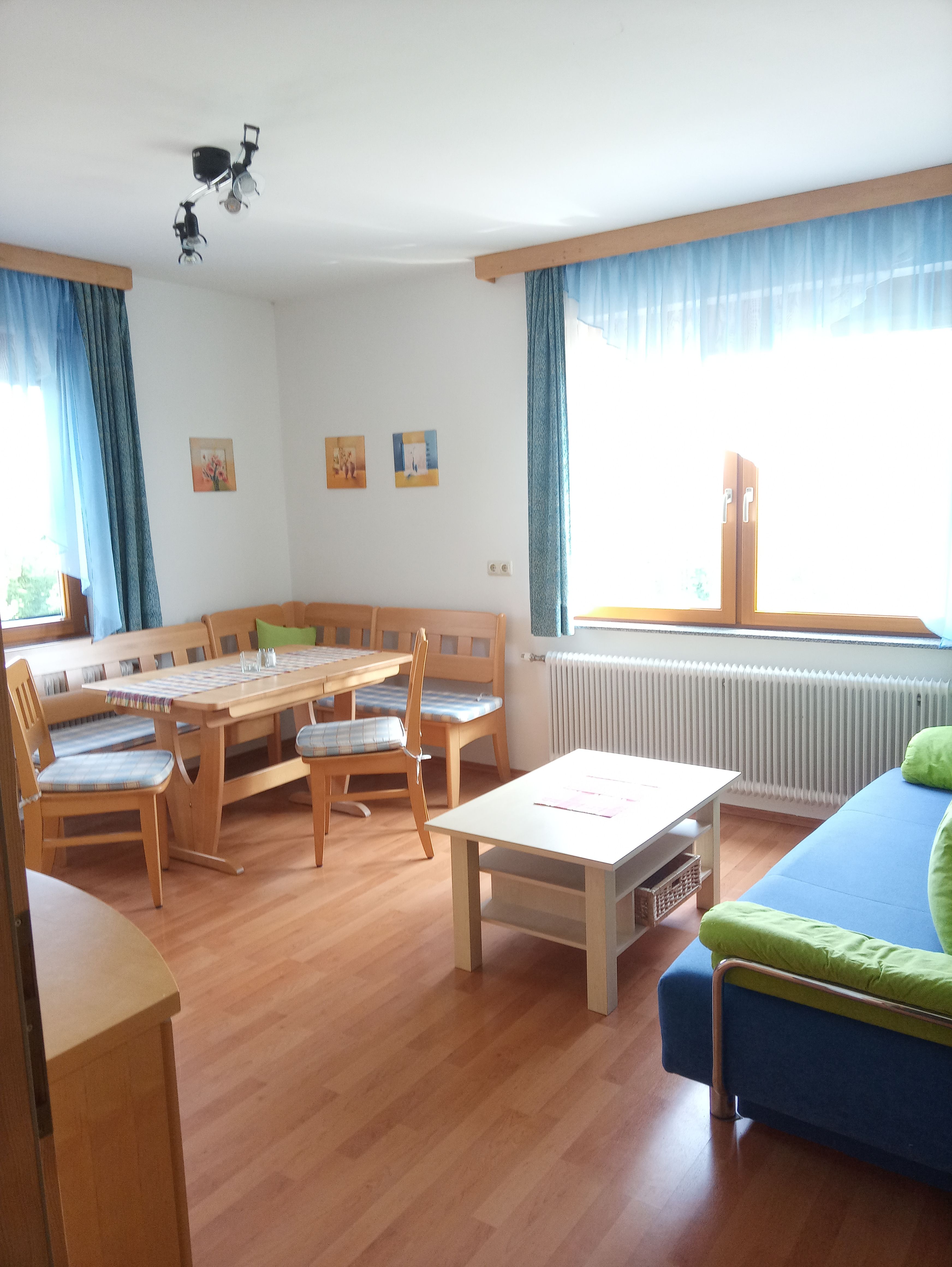 Világos nappali fabútorokkal, sarokpaddal, asztallal, kanapéval és nagy ablakokkal, kék függönyökkel.