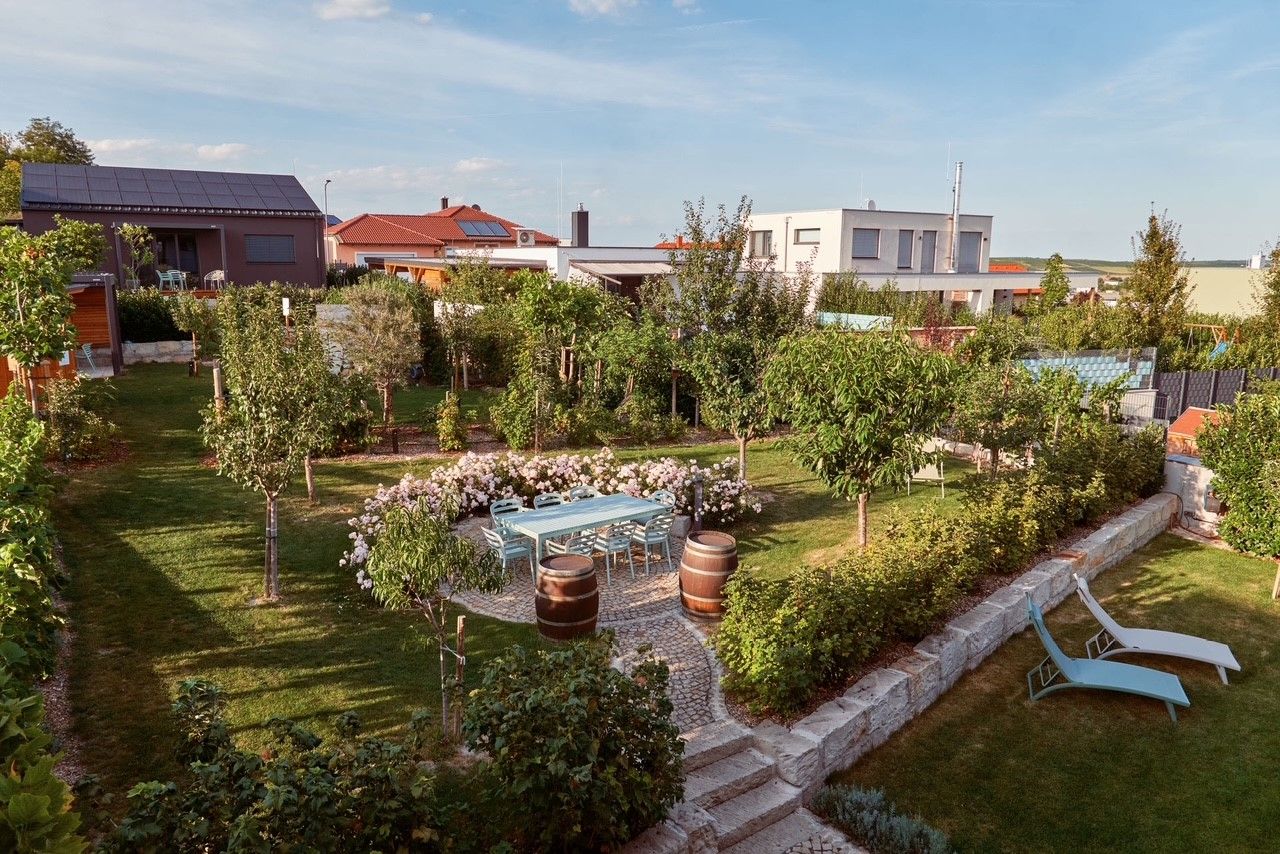 Gondozott kert fákkal, virágokkal, asztallal, székekkel és két nyugágydal. A háttérben modern házak láthatók.