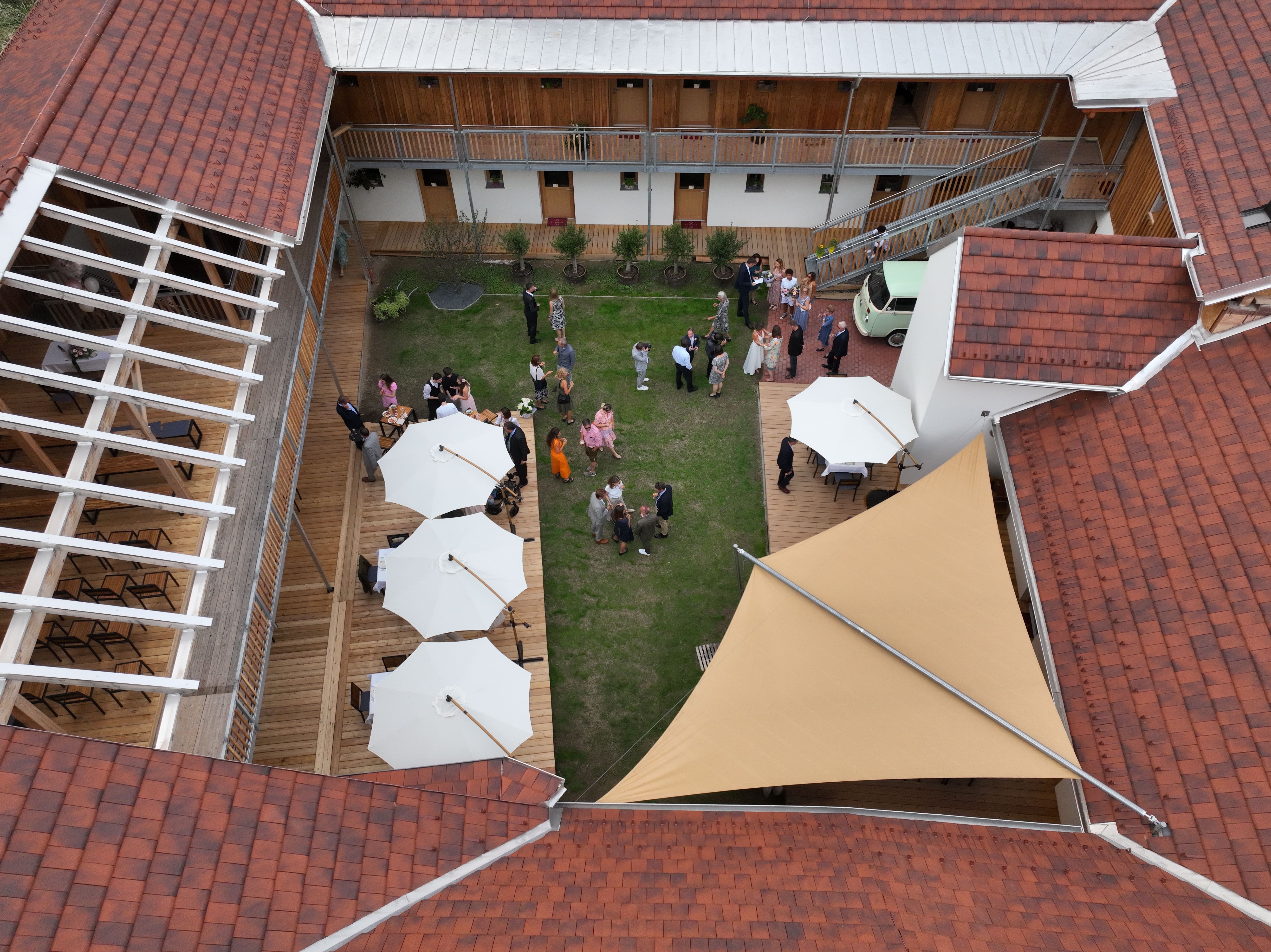 A belső udvar madártávlatból, zöld pázsittal, fehér napernyőkkel és vendégekkel egy fogadáson.