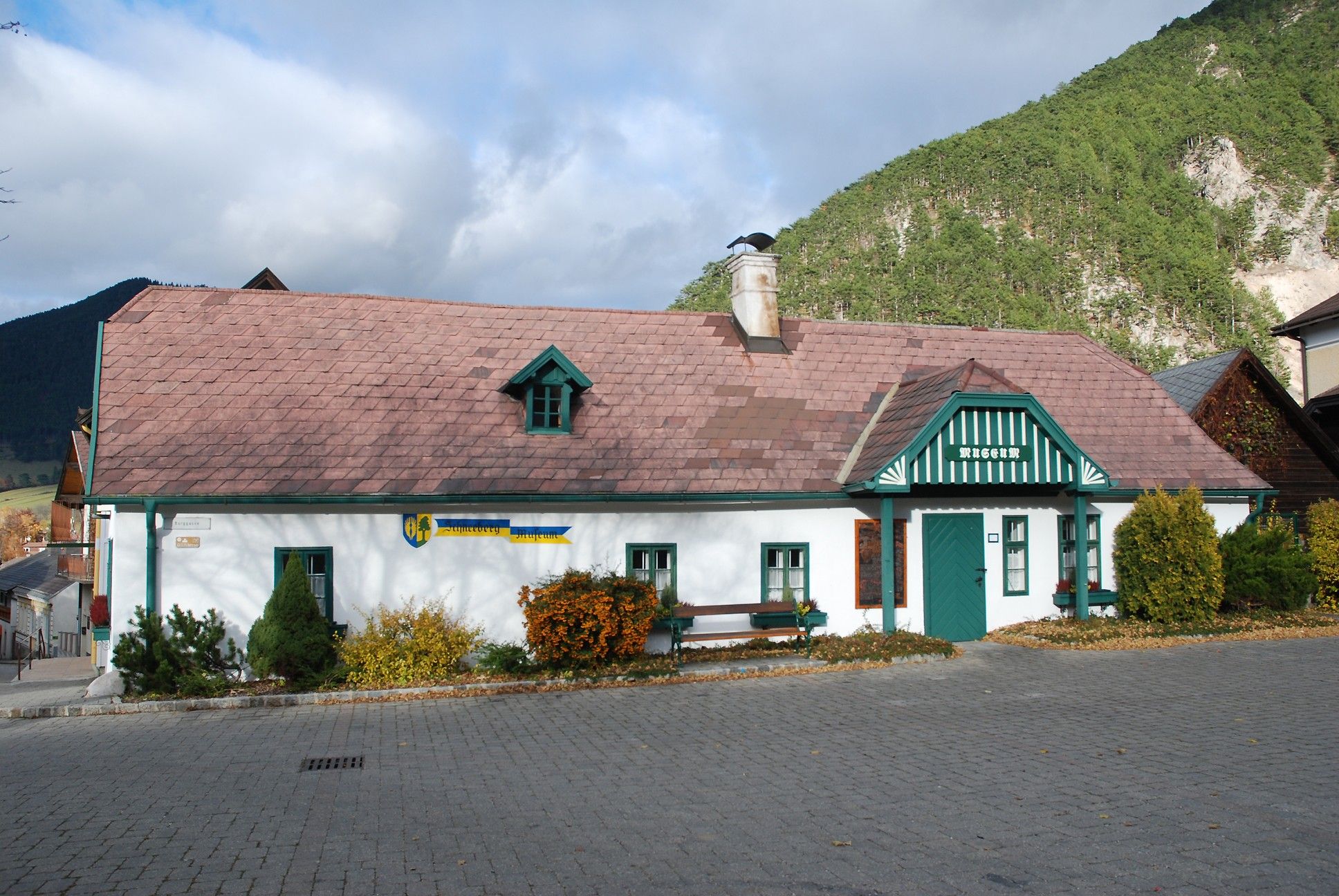 Schneeberg Museum Puchberg, egy hagyományos épület zöld tetővel és kéménynyel, hegyek és fák között.