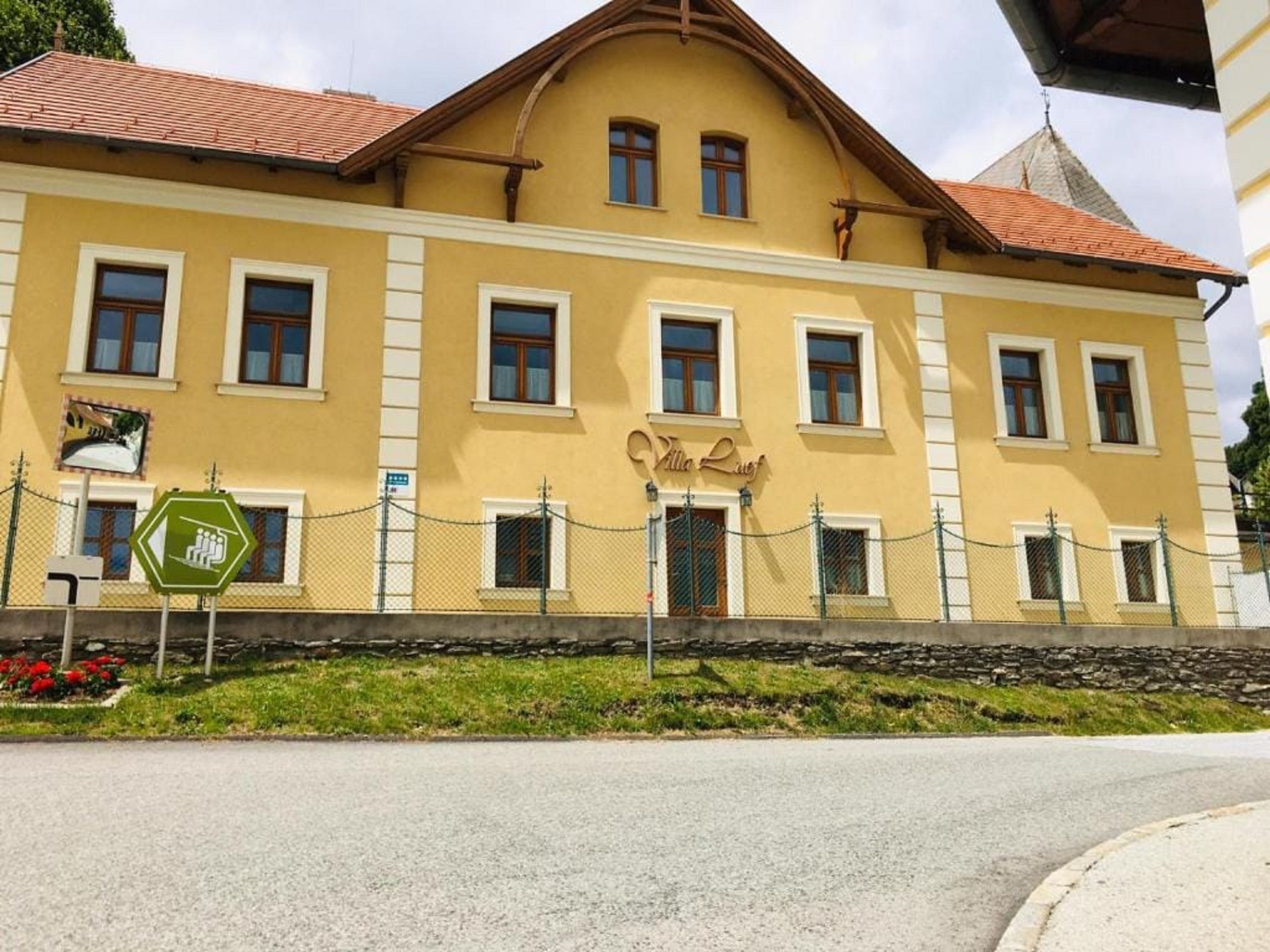 Sárga épület "Villa Luef" felirattal, kerítéssel körülvéve, előtérben út.
