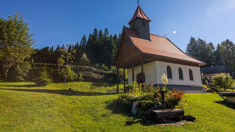 Piros tetejű kis kápolna egy zöld réten, fákkal és virágokkal körülvéve.