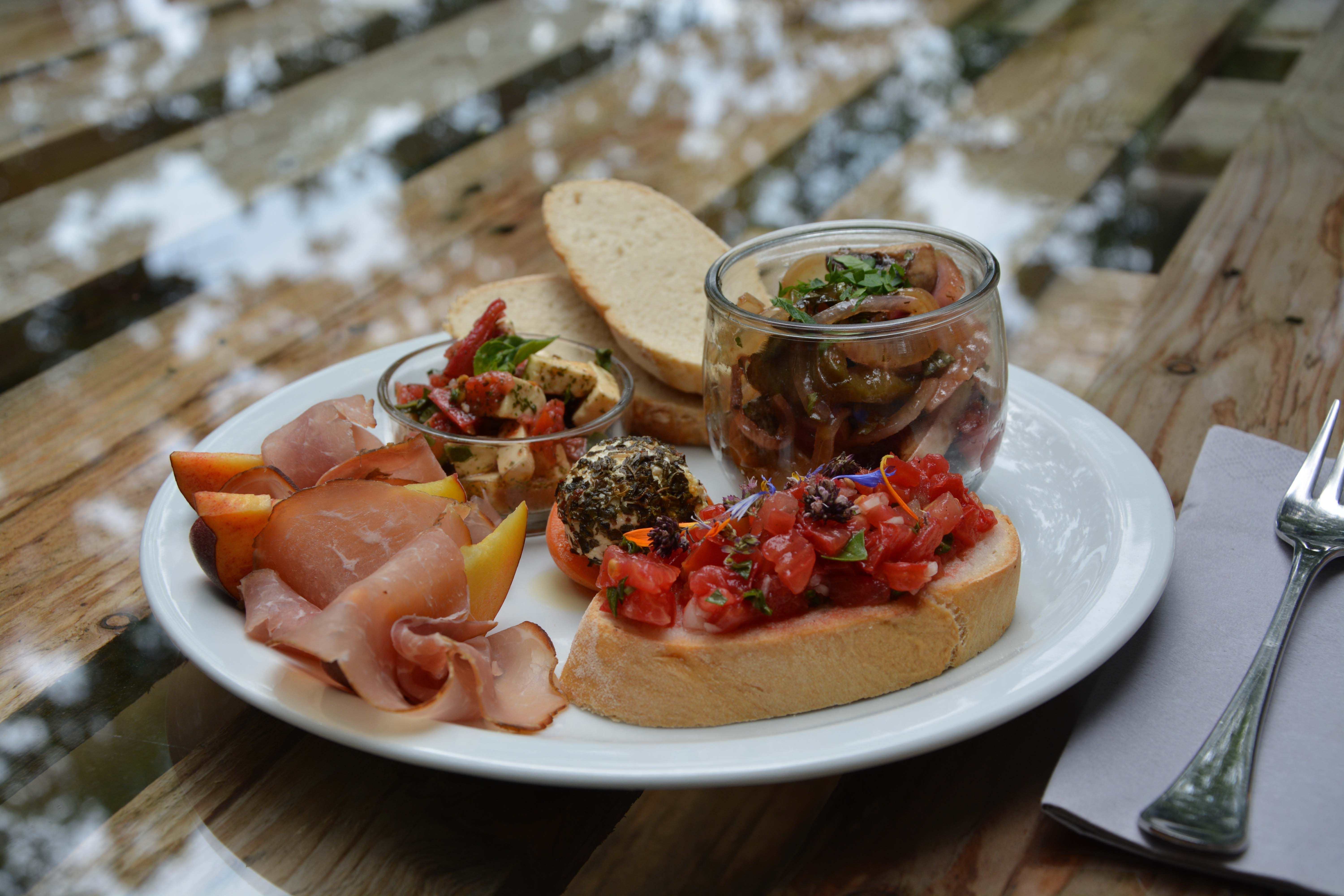 Egy tál kenyér, sonka, antipasti, bruschetta és gyümölcs egy faasztalon.