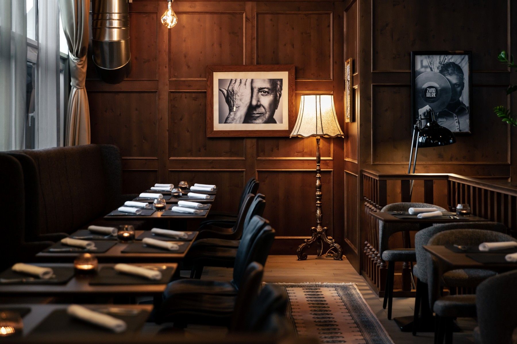 Hangulatos szoba fából készült falakkal, terített asztalokkal, állólámpával és fekete-fehér fényképekkel a falon.