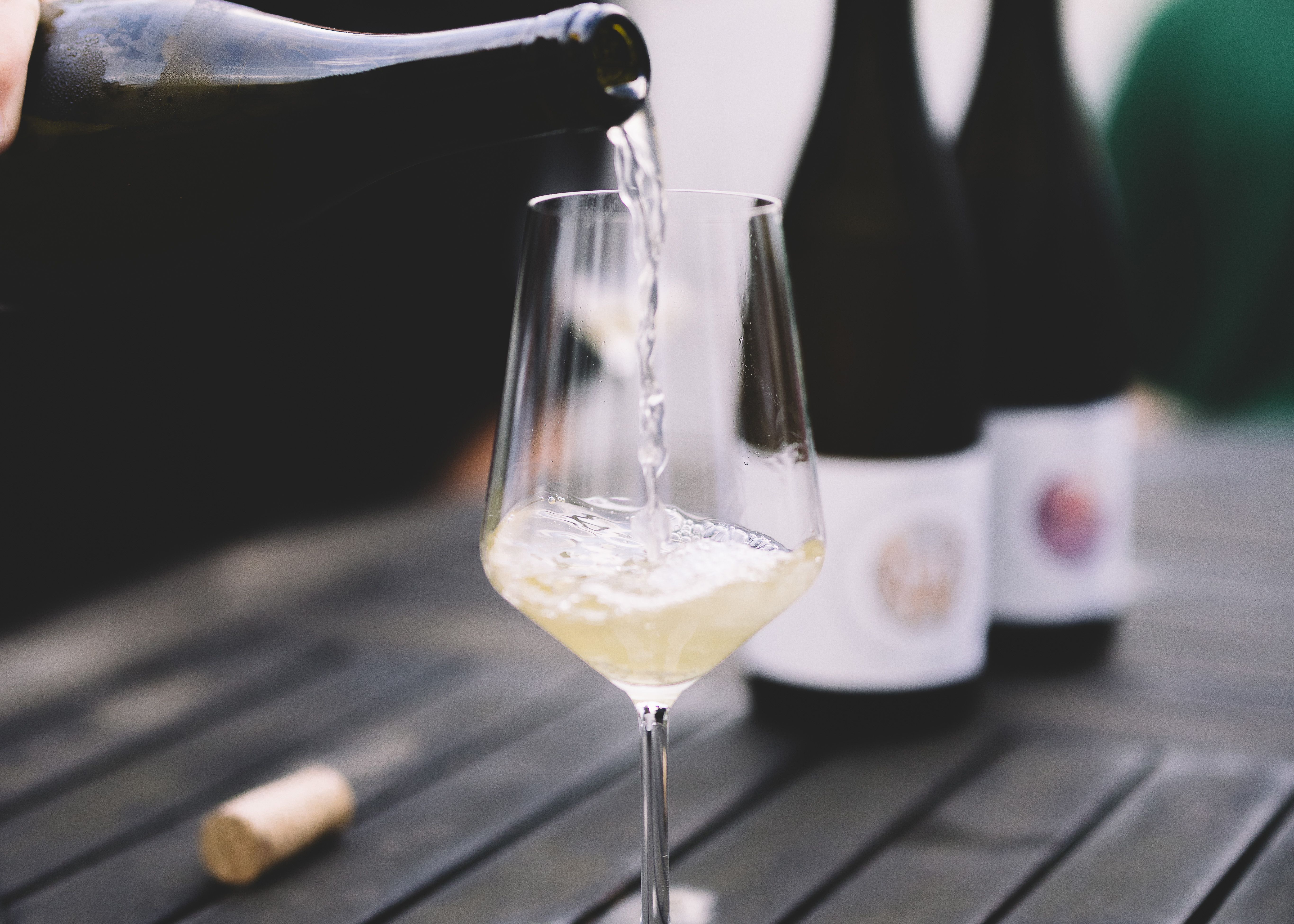 Egy pohár fehérbort töltenek egy asztalra, a háttérben két üveg borral.