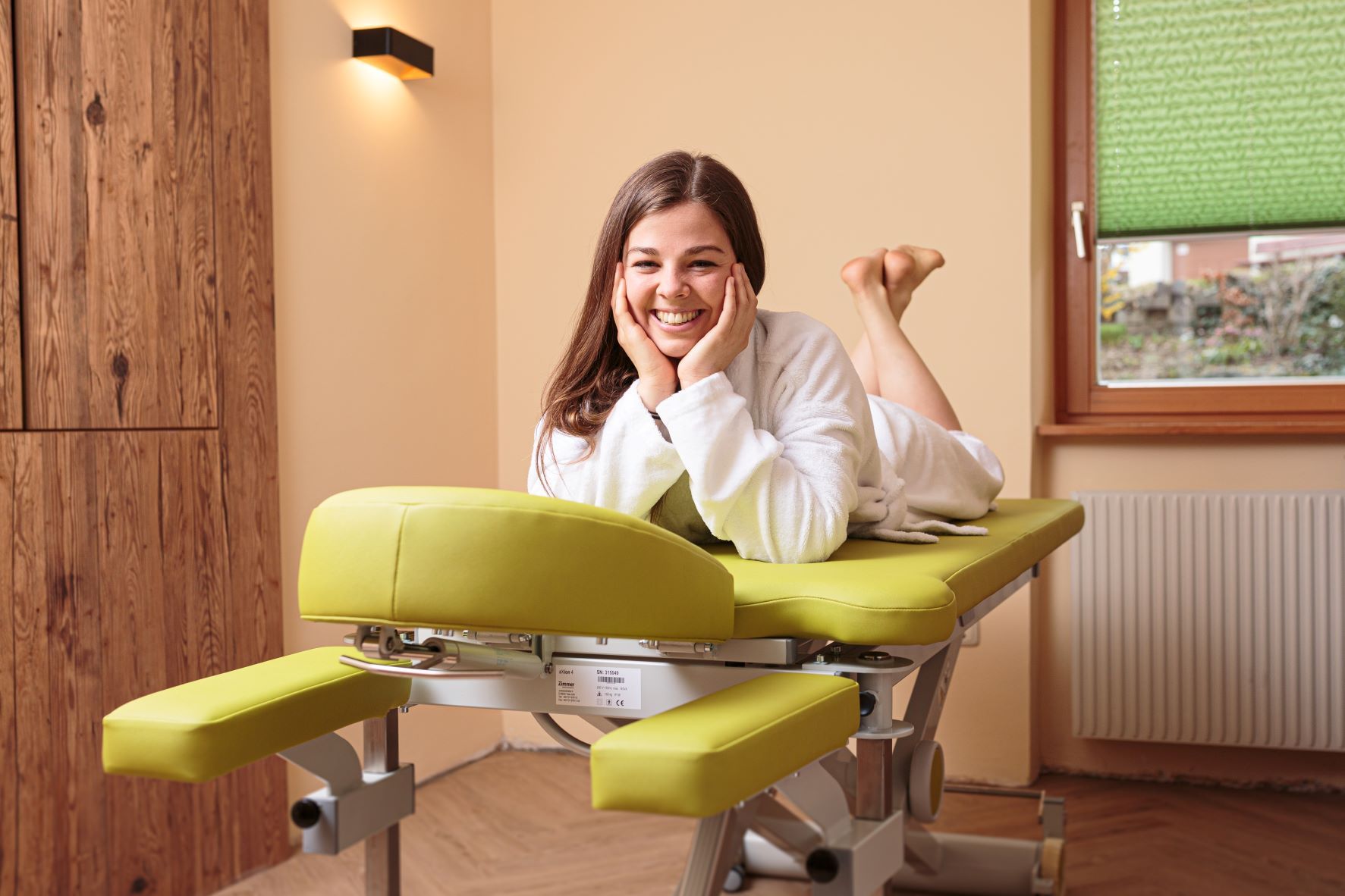 Fürdőköpenyes nő fekszik mosolyogva egy zöld masszázsasztalon egy wellness teremben.