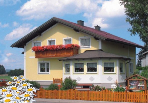 Sárga ház piros tetővel, az erkélyen virágládával, az előtérben fakerítéssel és százszorszépekkel körülvéve.
