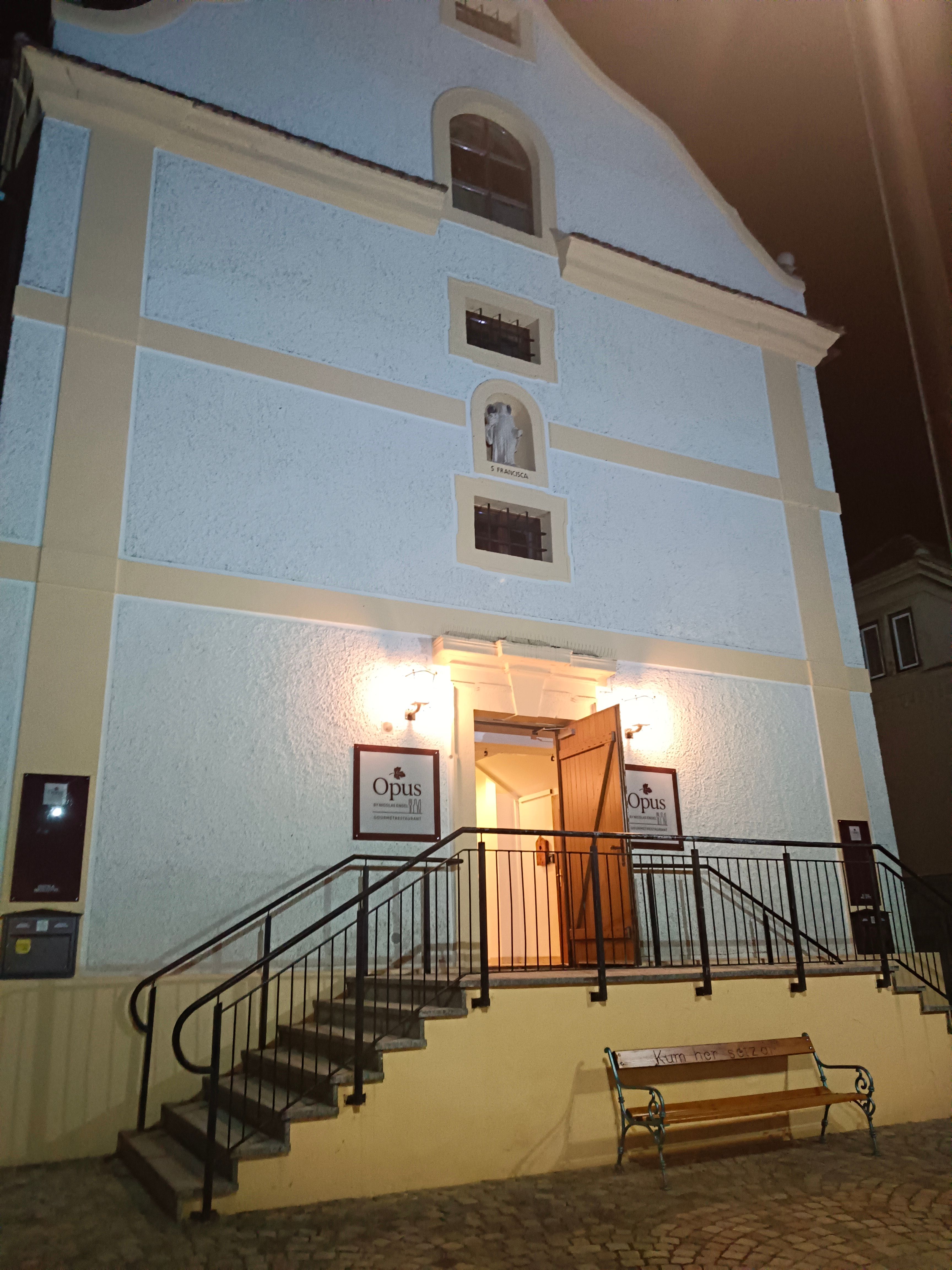 Éjjel megvilágított épület bejárata lépcsőkkel és nyitott ajtókkal.