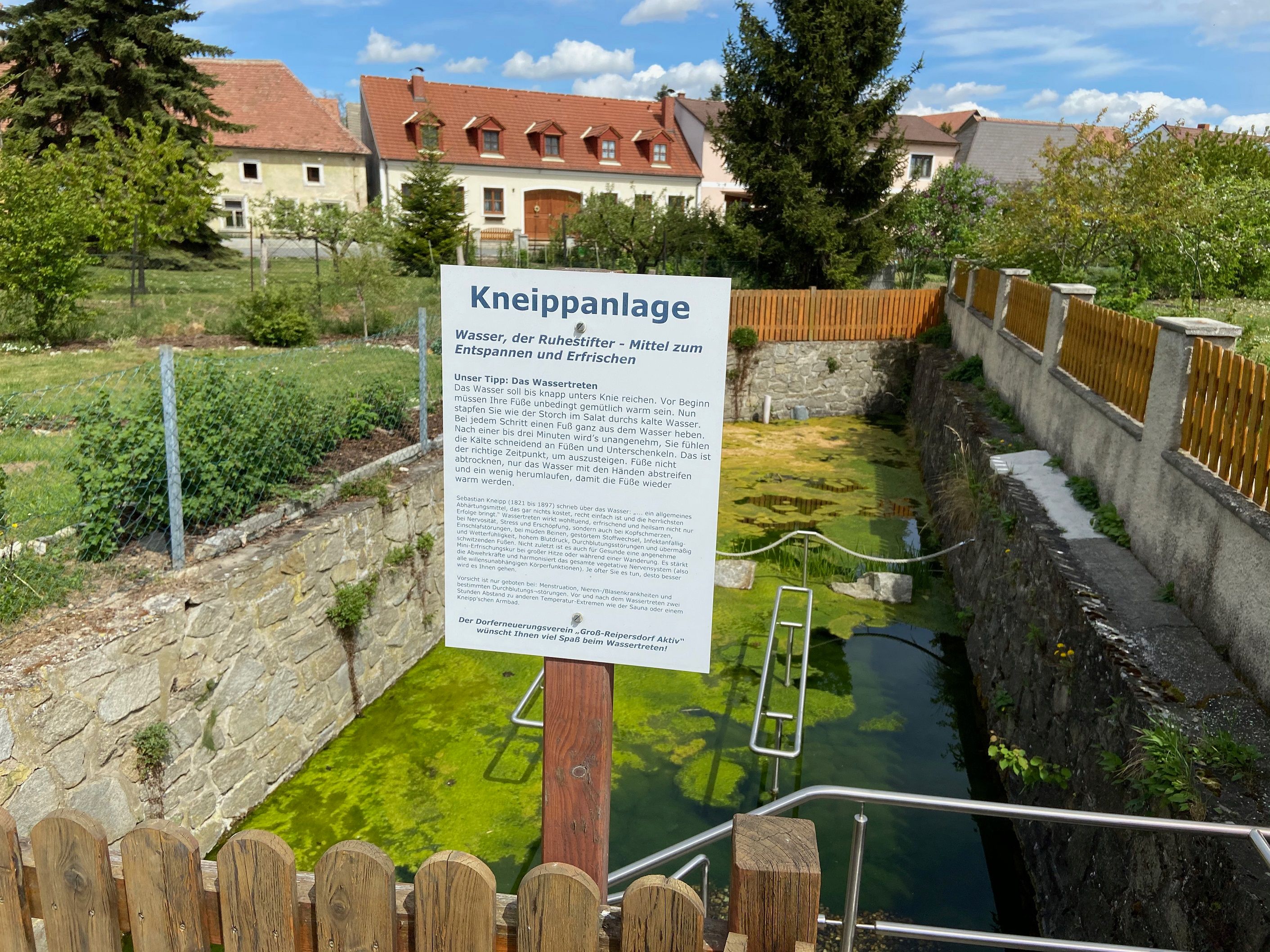 Kneipp-létesítmény információs táblával és vízmedencével, kerítéssel körülvéve, a háttérben házakkal.