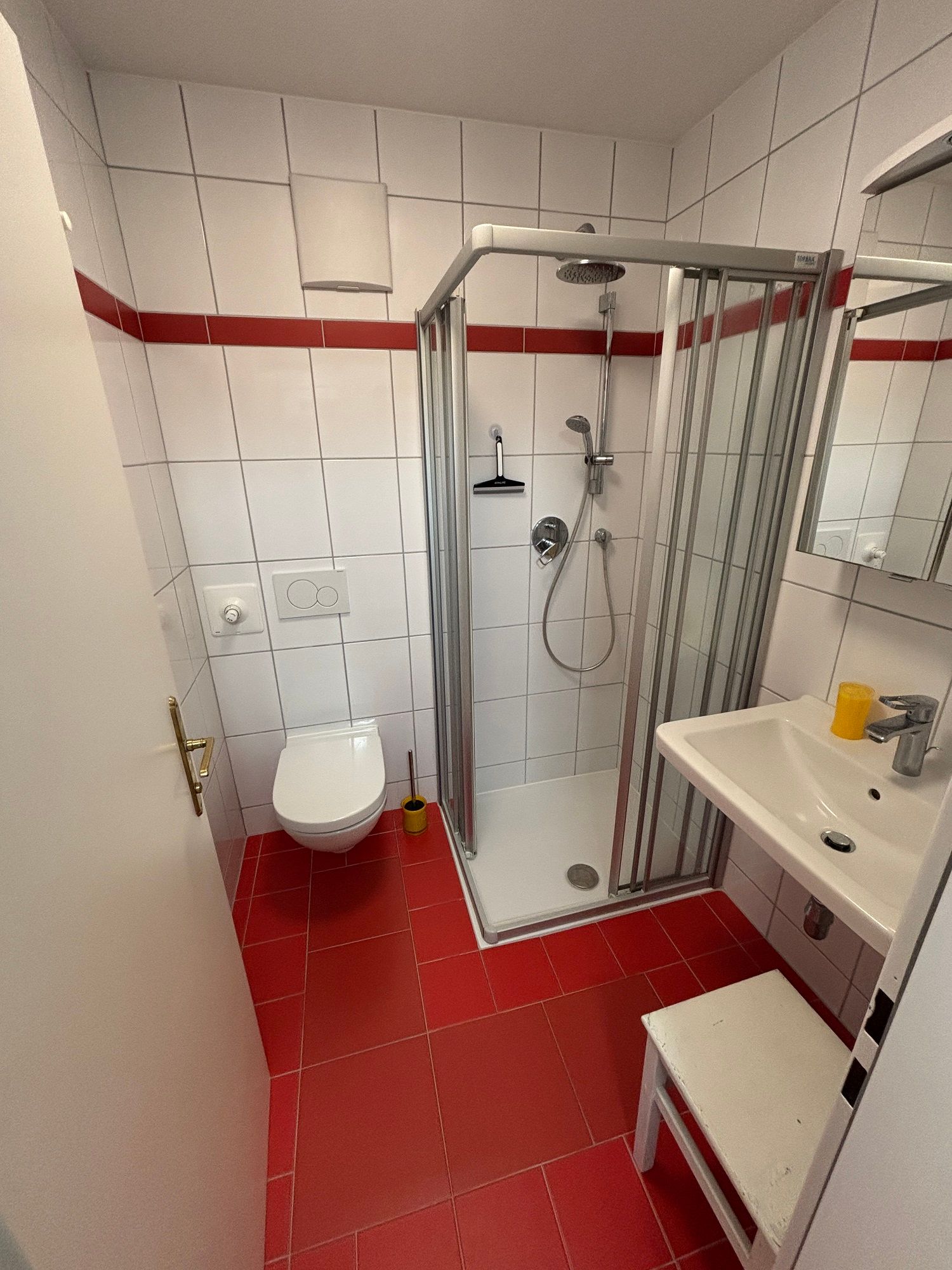 Fürdőszoba piros csempézett padlóval, fehér csempézett falakkal, zuhanyzóval, mosdóval és WC-vel.