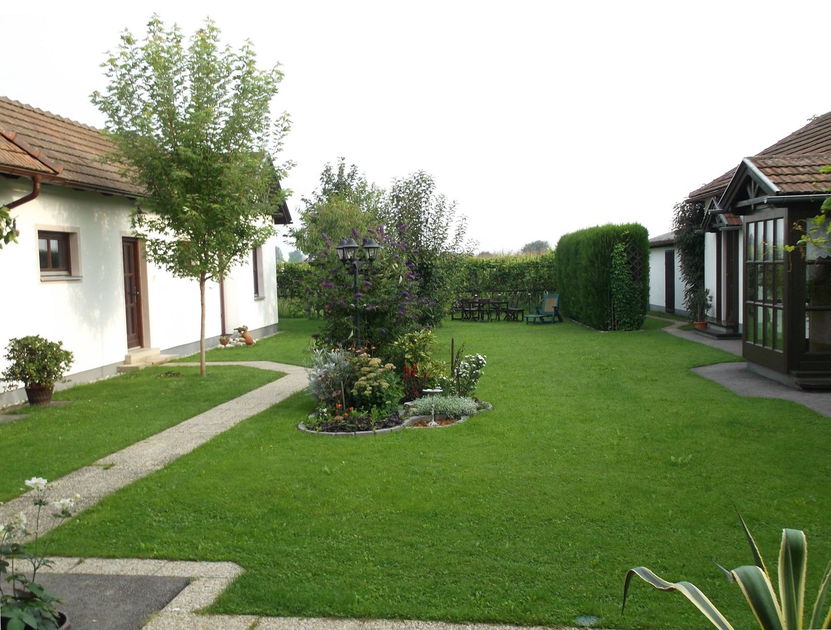 Nagyon gondozott kert, kilátással a bokrokra és fákra, a háttérben az épület egy részével.