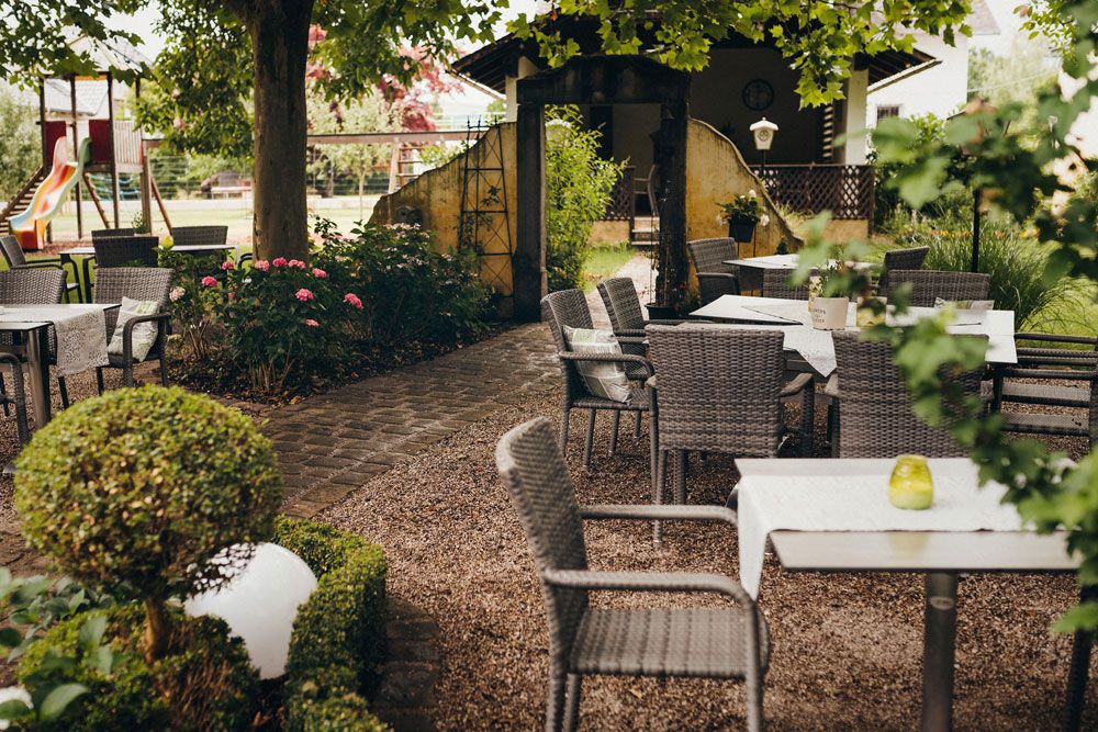 Hangulatos szabadtéri terület asztalokkal és székekkel a fák alatt, növényekkel körülvéve, a háttérben játszótérrel.