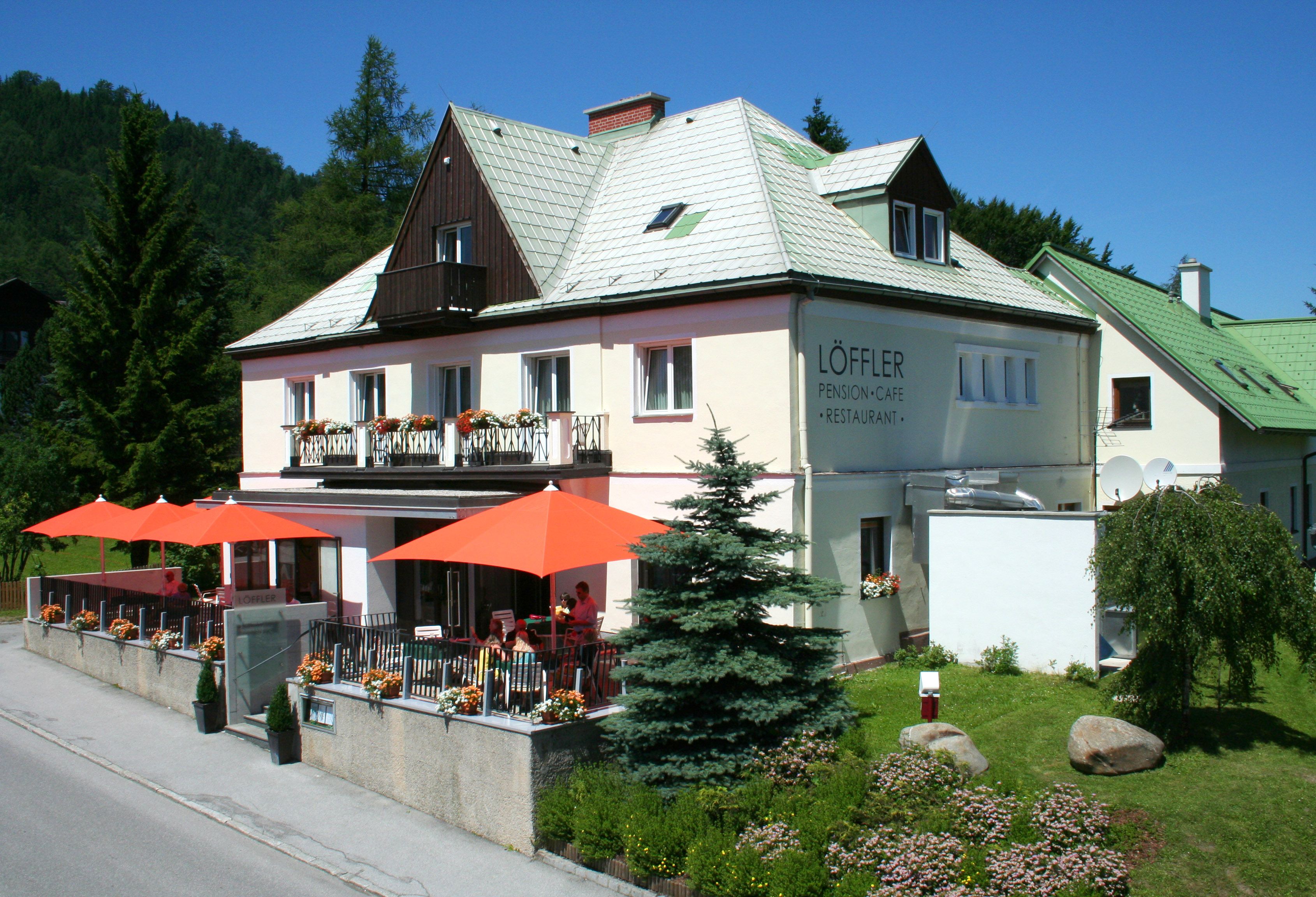 A "Löffler Panzió-Café-Restaurant" feliratú kétszintes épület, fákkal és virágokkal körülvéve, a teraszon narancssárga napernyőkkel.