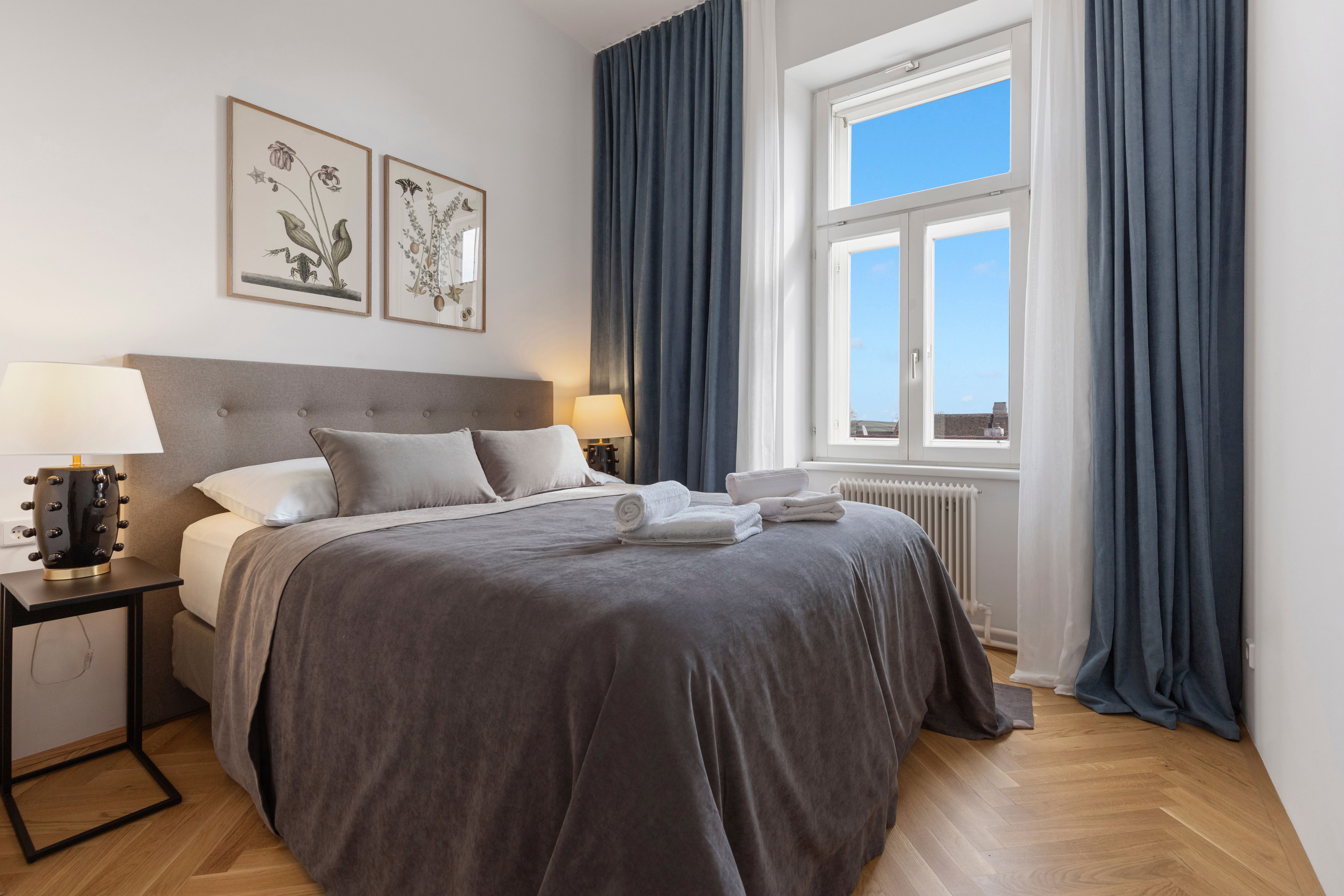 Elegáns hálószoba nagy ággyal, szürke ágyneművel, két éjjeliszekrénnyel és lámpával, valamint kék függönnyel ellátott ablakkal.