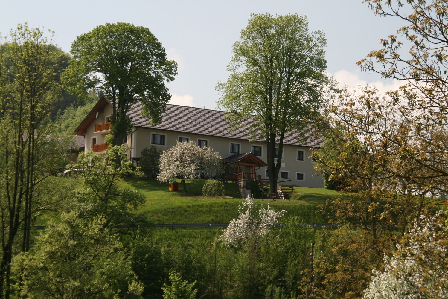 Egy farm egy zöld dombon, fákkal és virágzó bokrokkal körülvéve.