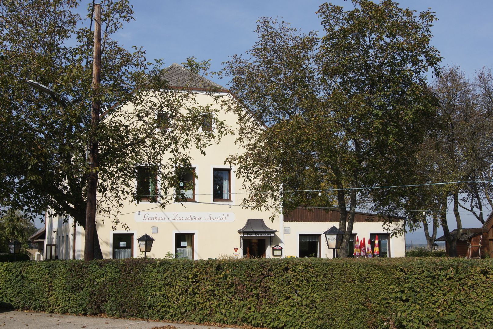 A "Gasthaus Zur schönen Aussicht" feliratú sárga vendéglő, fákkal és sövényekkel körülvéve.