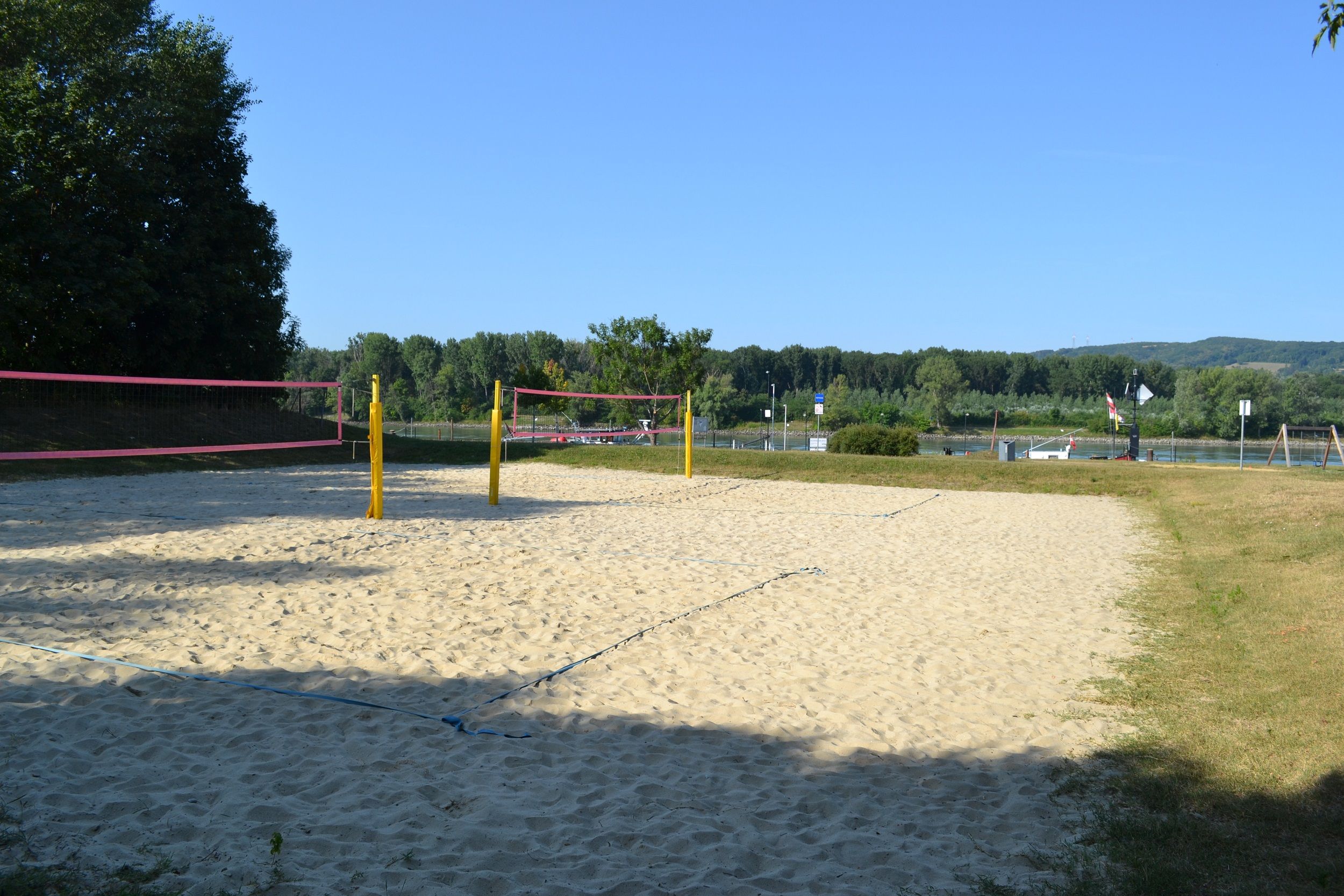 Egy üres strandröplabdapálya sárga oszlopokkal és rózsaszín hálóval, a háttérben fák és egy folyó.