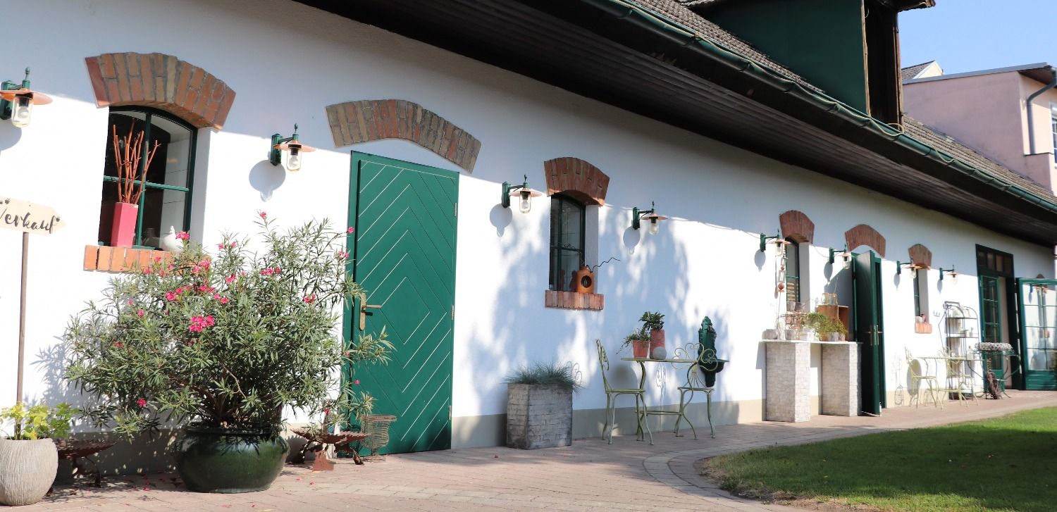 Fehér épület zöld ajtókkal és ablakokkal, növényekkel és kerti bútorokkal körülvéve.