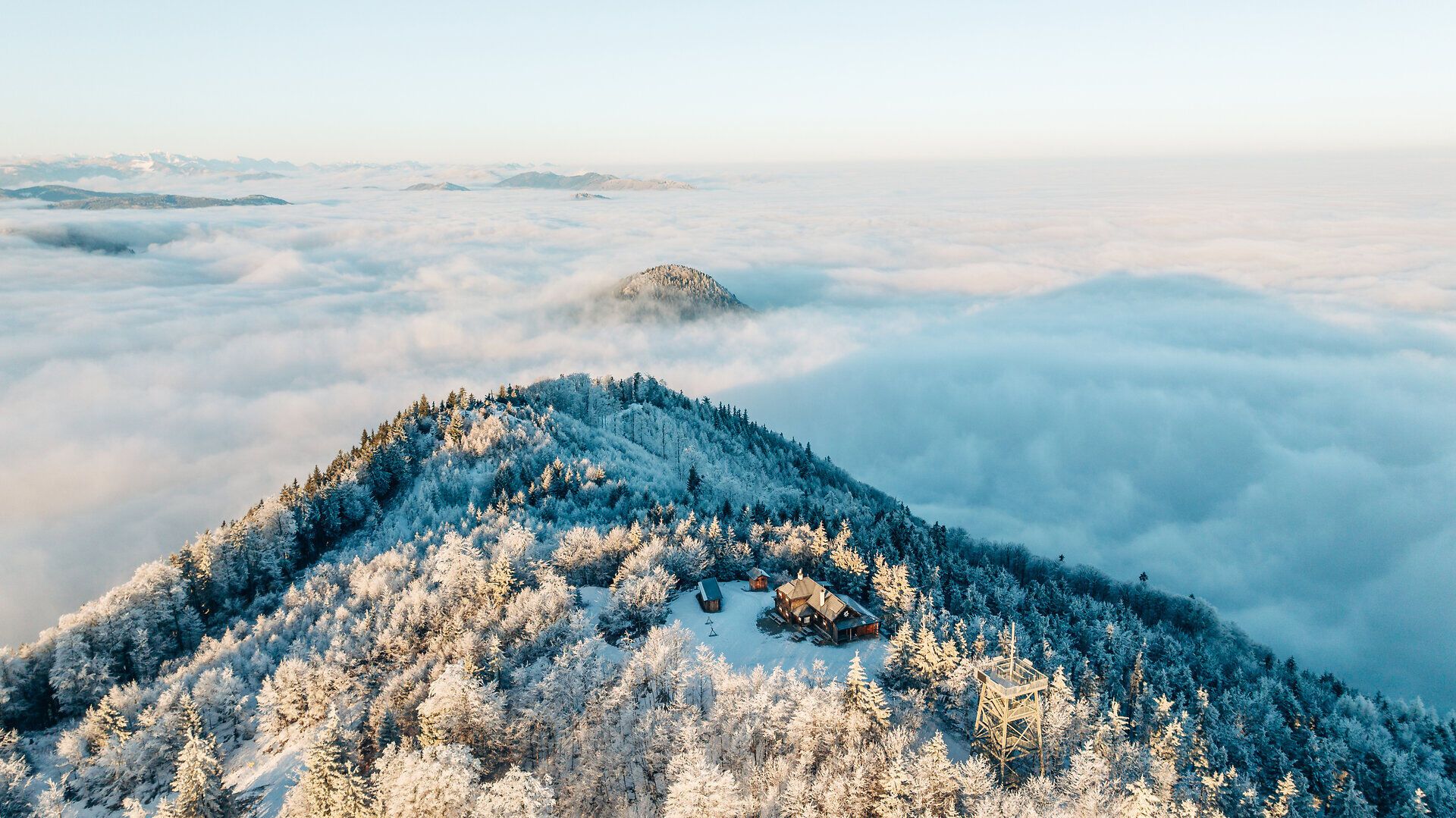 Verschneiter Berggipfel mit Hütte und Aussichtsturm über einem weiten Nebelmeer.
